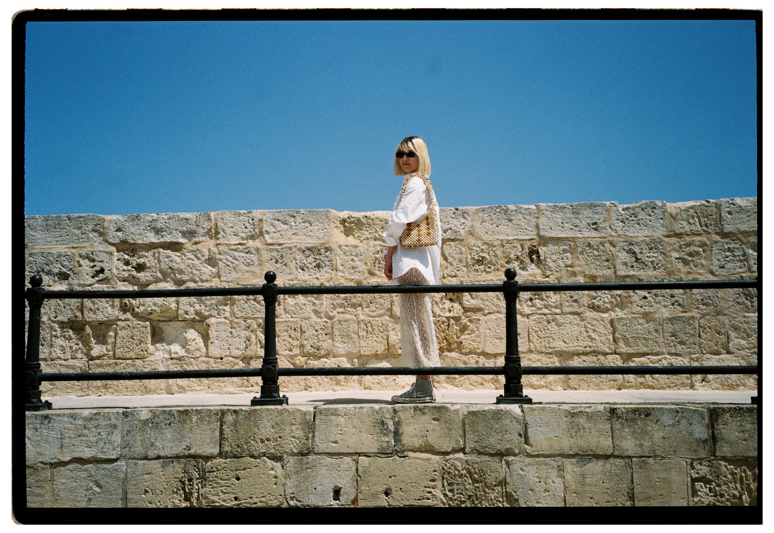 A woman with blonde hair and sunglasses standing on a stone walkway behind a black metal fence, with a stone wall and a clear blue sky in the background. with her beaded bag