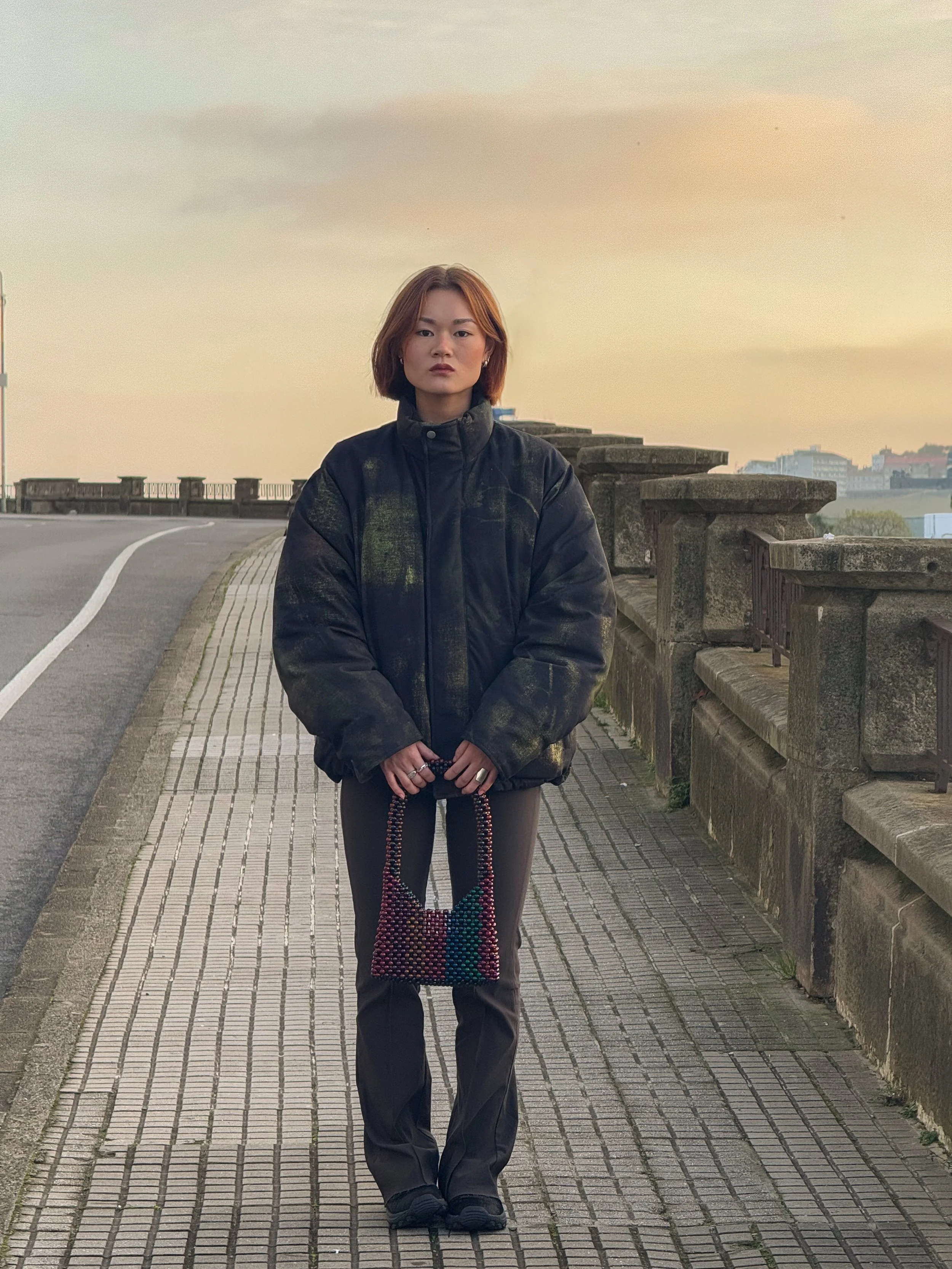 A woman with short brown hair standing on a sidewalk during sunset, wearing a black jacket, brown pants, and holding a colorful beaded handbag.
