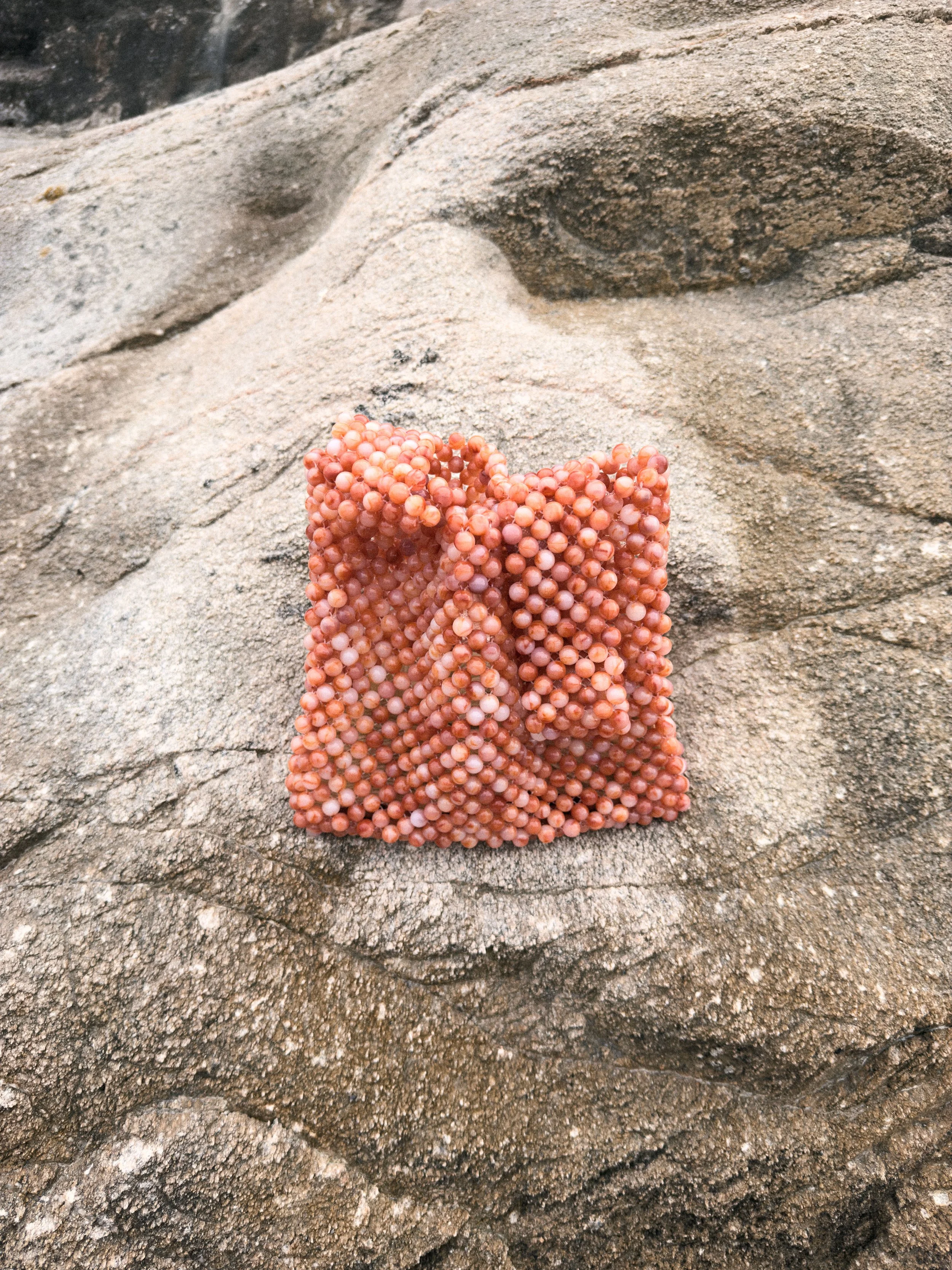 Pinkish-orange beaded clutch purse resting on sandy, rocky surface.