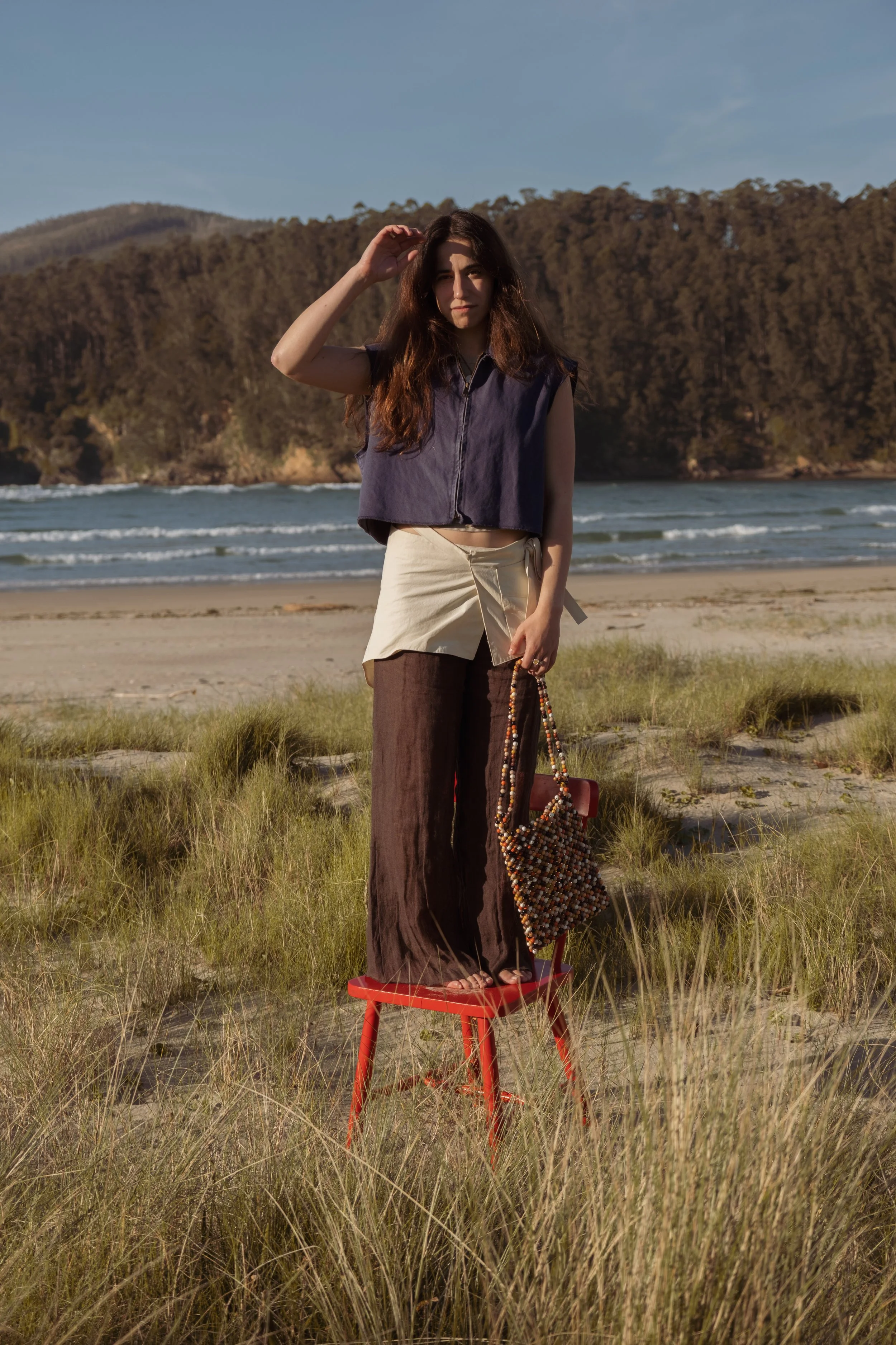 A woman standing on a red chair on a beach, holding a beaded bag, with an ocean and forested hills in the background.