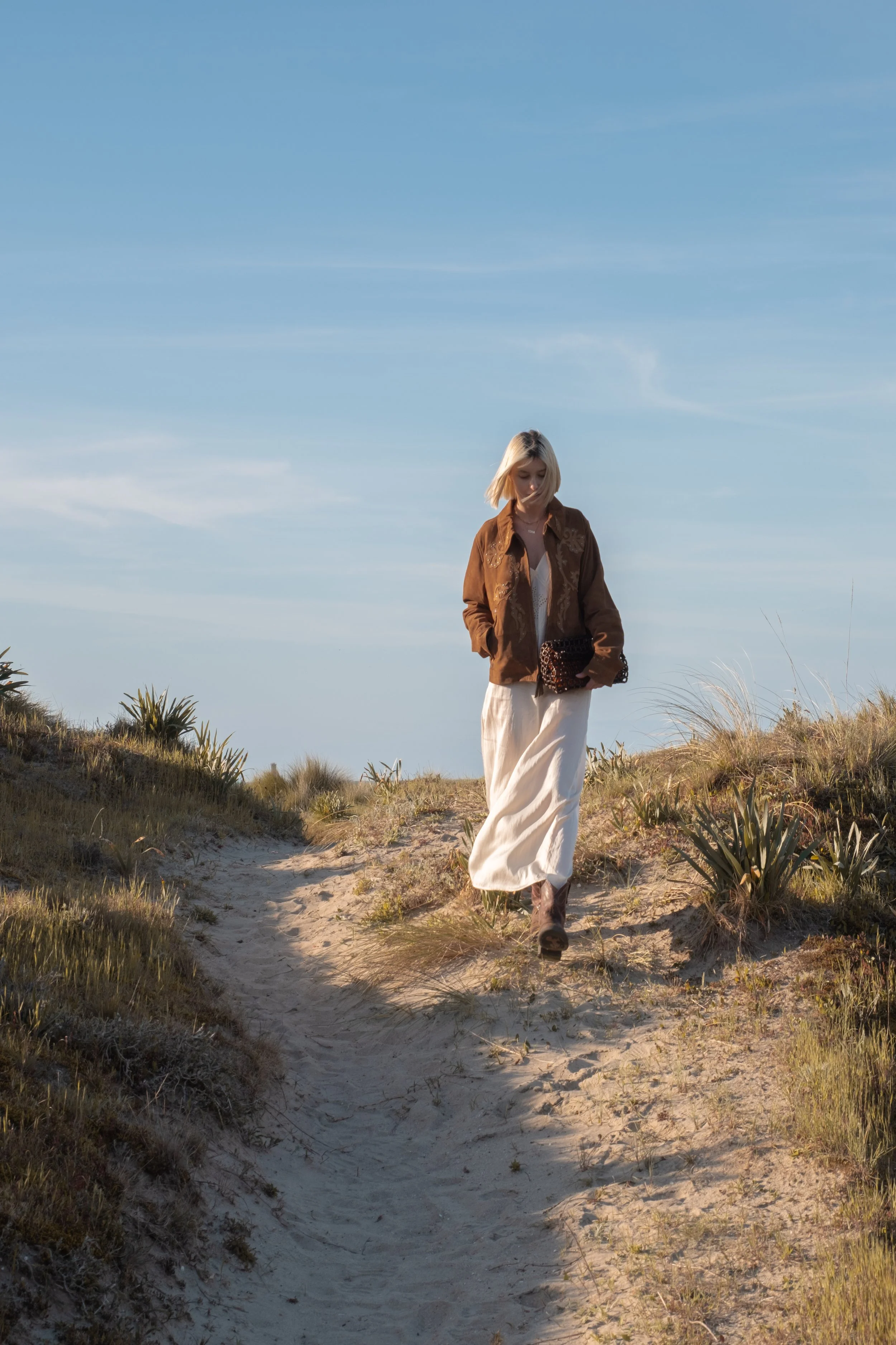 A woman walking along a sandy trail with grass and plants, under a clear blue sky.