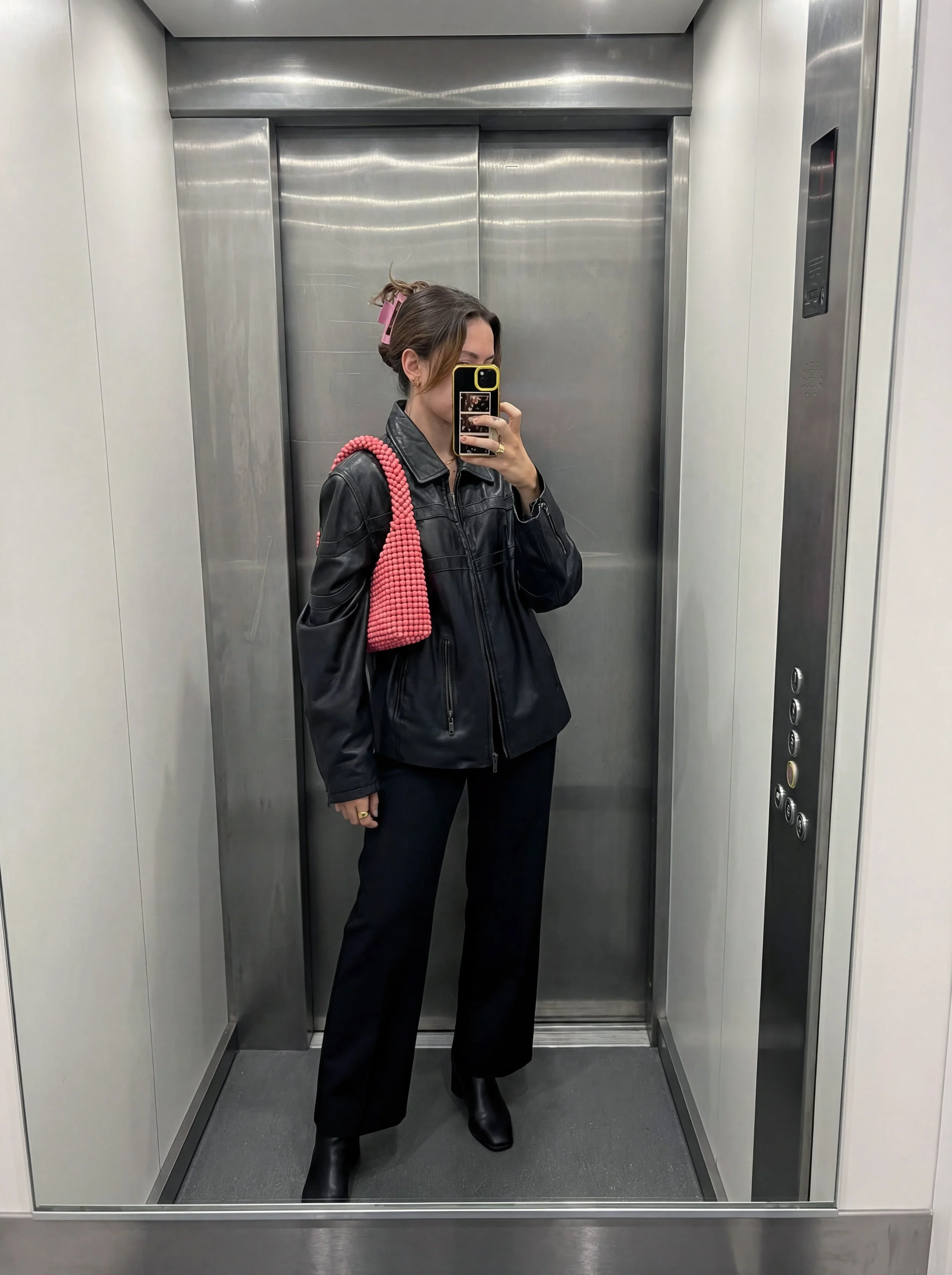 A woman taking a mirror selfie inside an elevator. She is wearing a black leather jacket, black pants, and black boots. She has a pink hair clip and is carrying a pink, beaded bag over her shoulder. She is holding her phone with a black and yellow case.