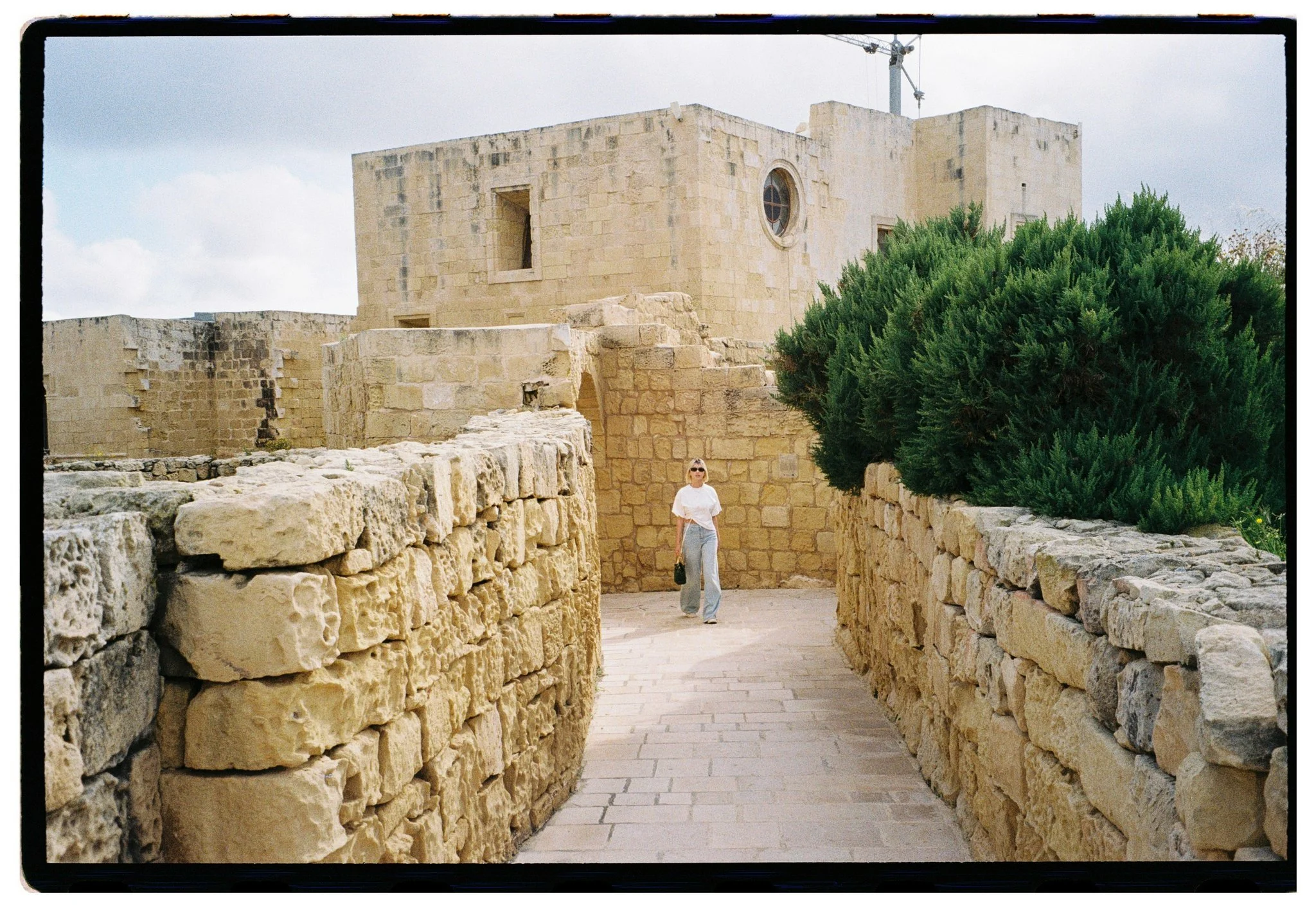 A woman walking through a historic stone fortress with beige limestone walls and greenery, under a cloudy sky.