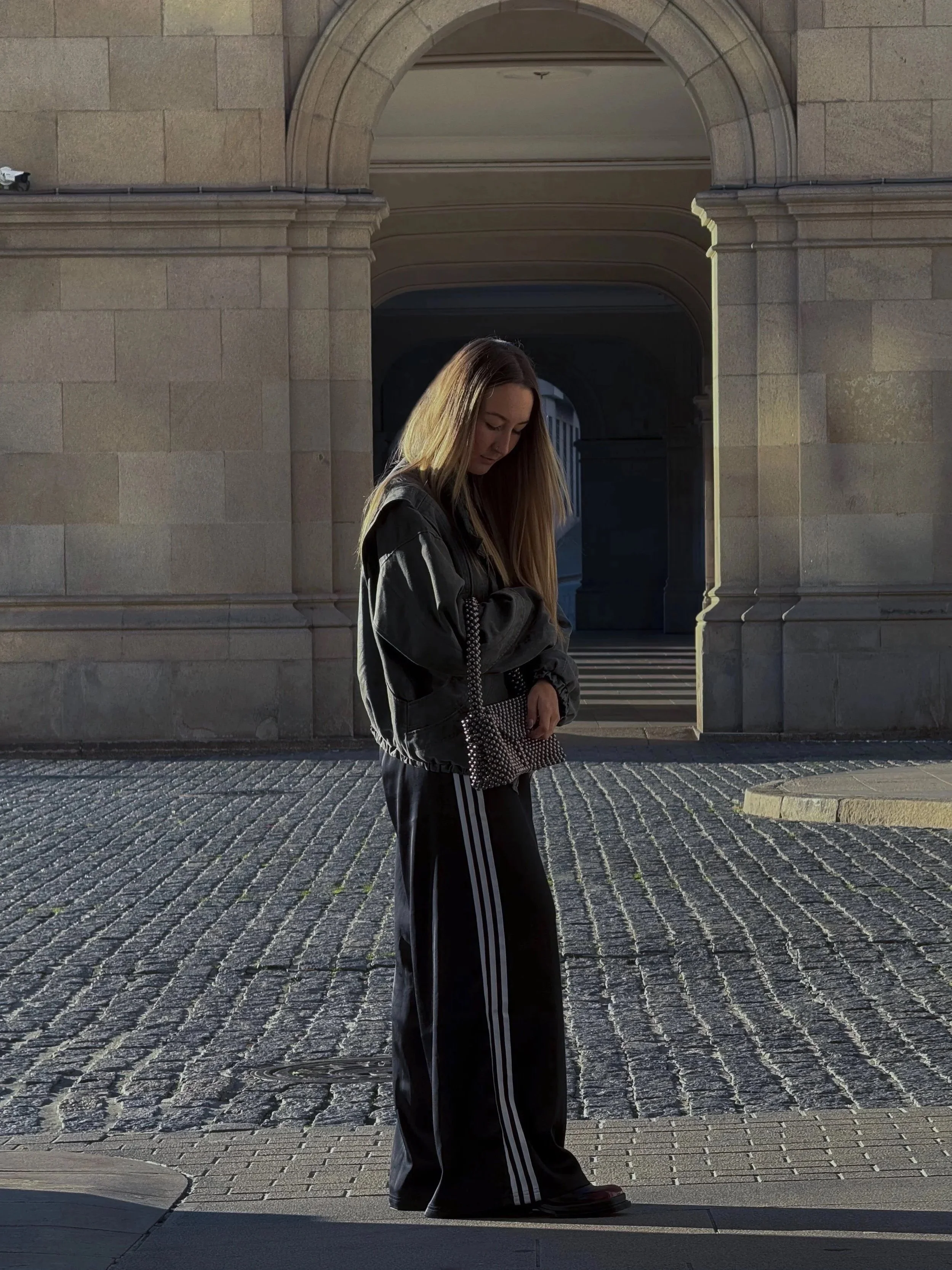 A young woman with long blonde hair standing on a cobblestone street in front of a stone building with an arched entrance, wearing a black jacket and black Adidas pants with white stripes, carrying a studded purse.