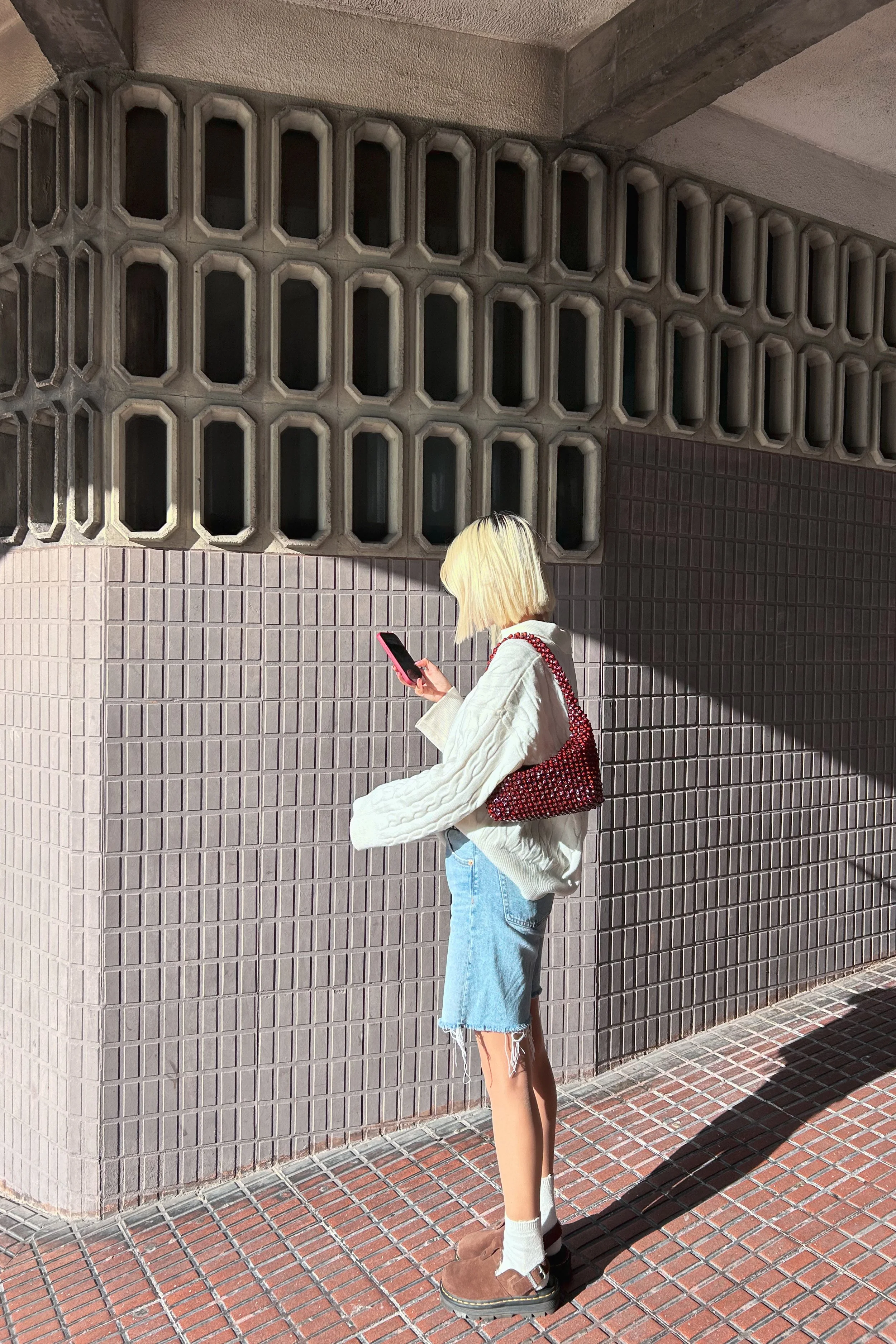 A young woman with blonde hair and wearing a beige sweater, denim shorts, and brown shoes is standing on a brick sidewalk, looking at her smartphone. She has a red patterned bag slung over her shoulder. The background features a concrete wall with a distinctive geometric window design and a shadow cast on the ground from the wall.