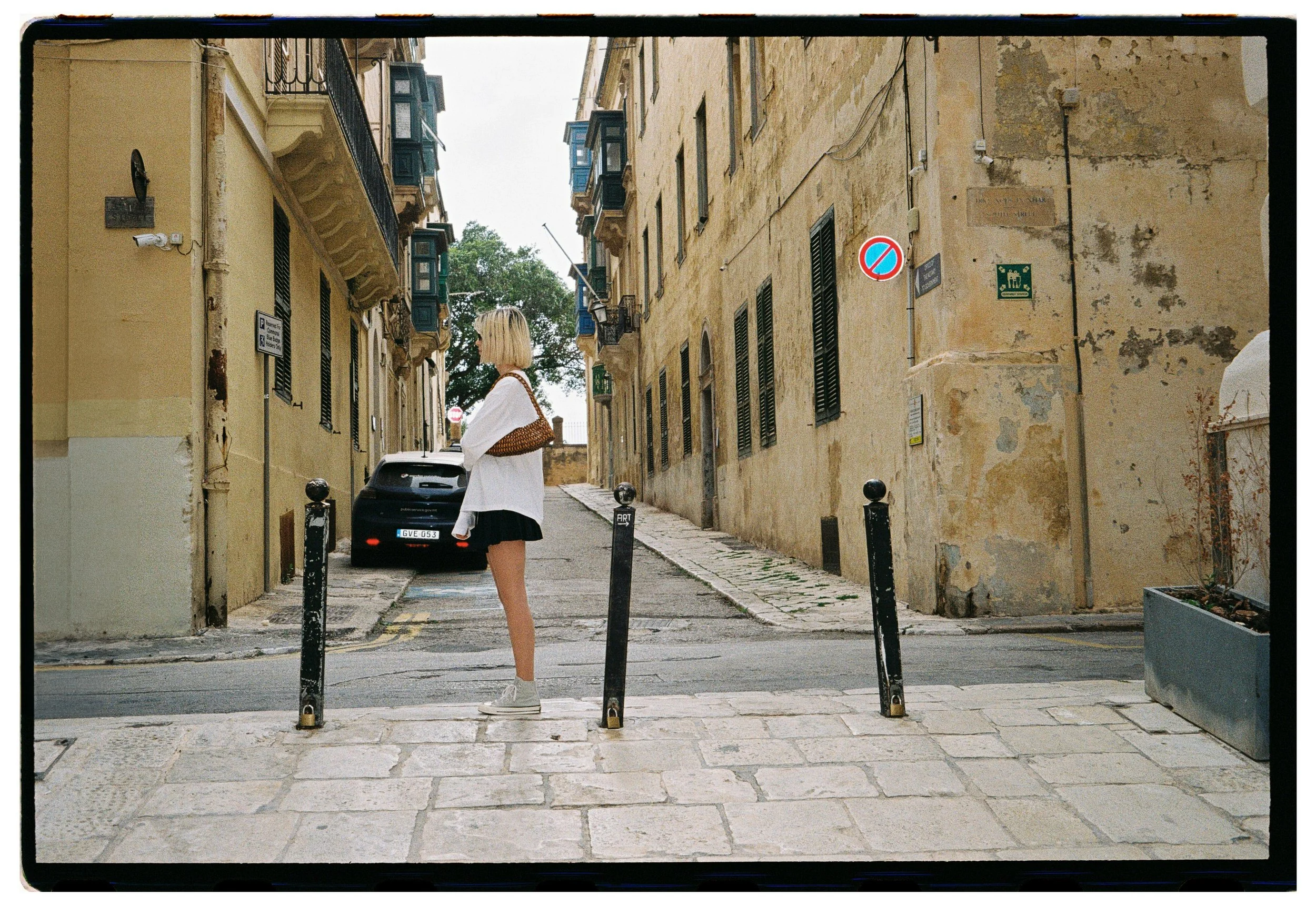 A woman with blonde hair wearing a white jacket, black skirt, and white sneakers stands on a cobblestone crosswalk in an old European street, surrounded by yellow weathered buildings with blue balconies and a parked car behind her. holding her beaded