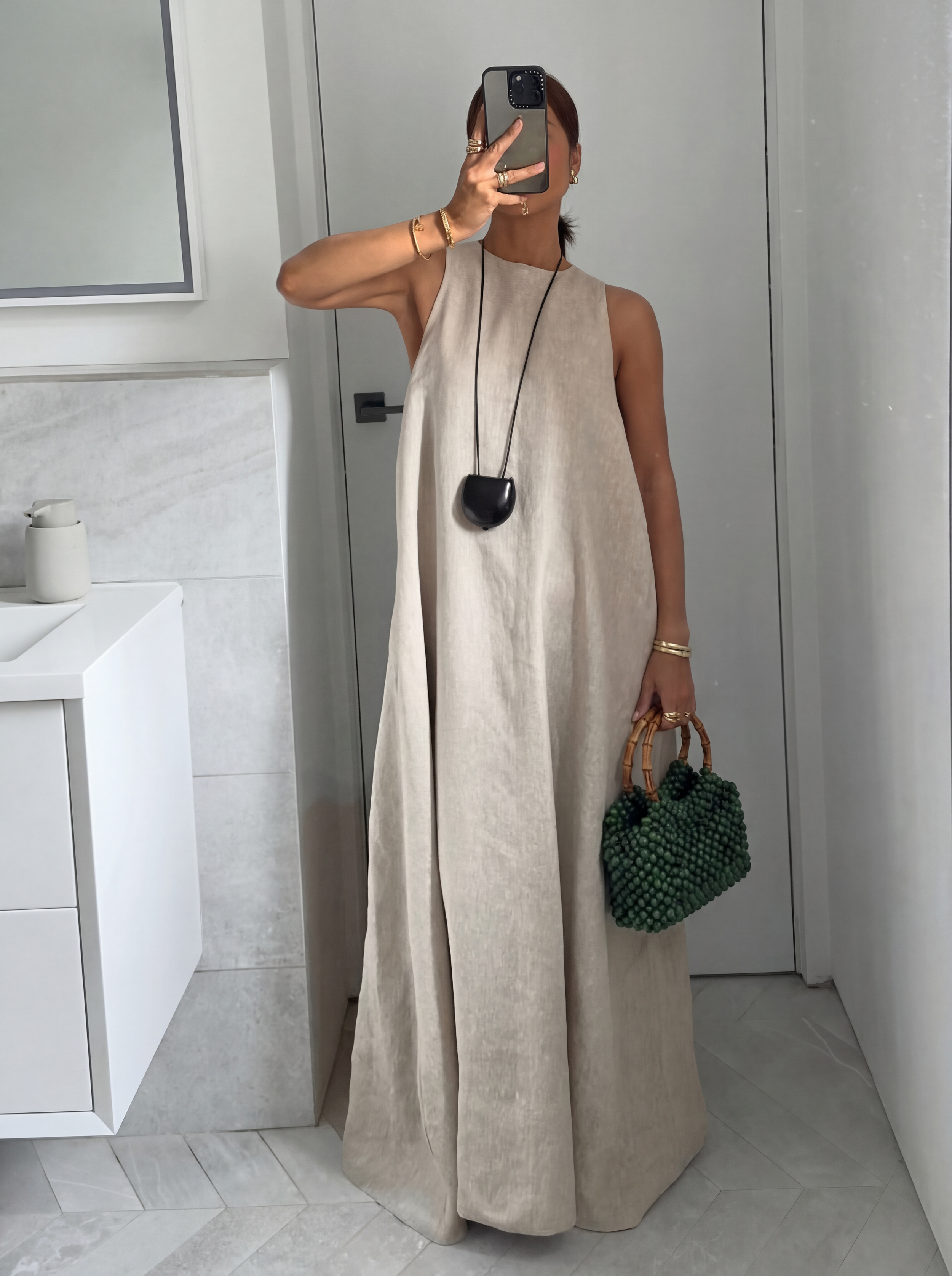 Woman in a beige sleeveless dress taking a mirror selfie, holding a green beaded handbag with bamboo handles, and wearing gold jewelry.