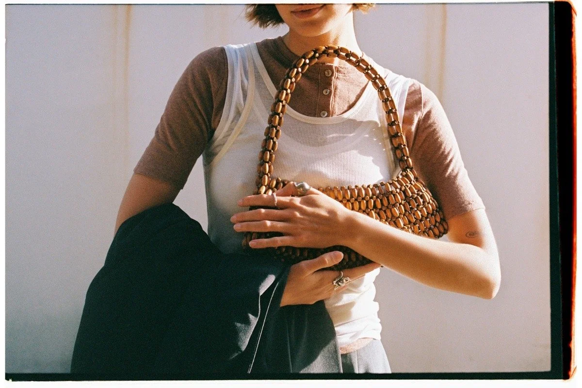 Person holding a wooden beaded handbag in front of their chest, wearing a layered short-sleeve shirt and necklace, with a dark jacket draped over their arm.
