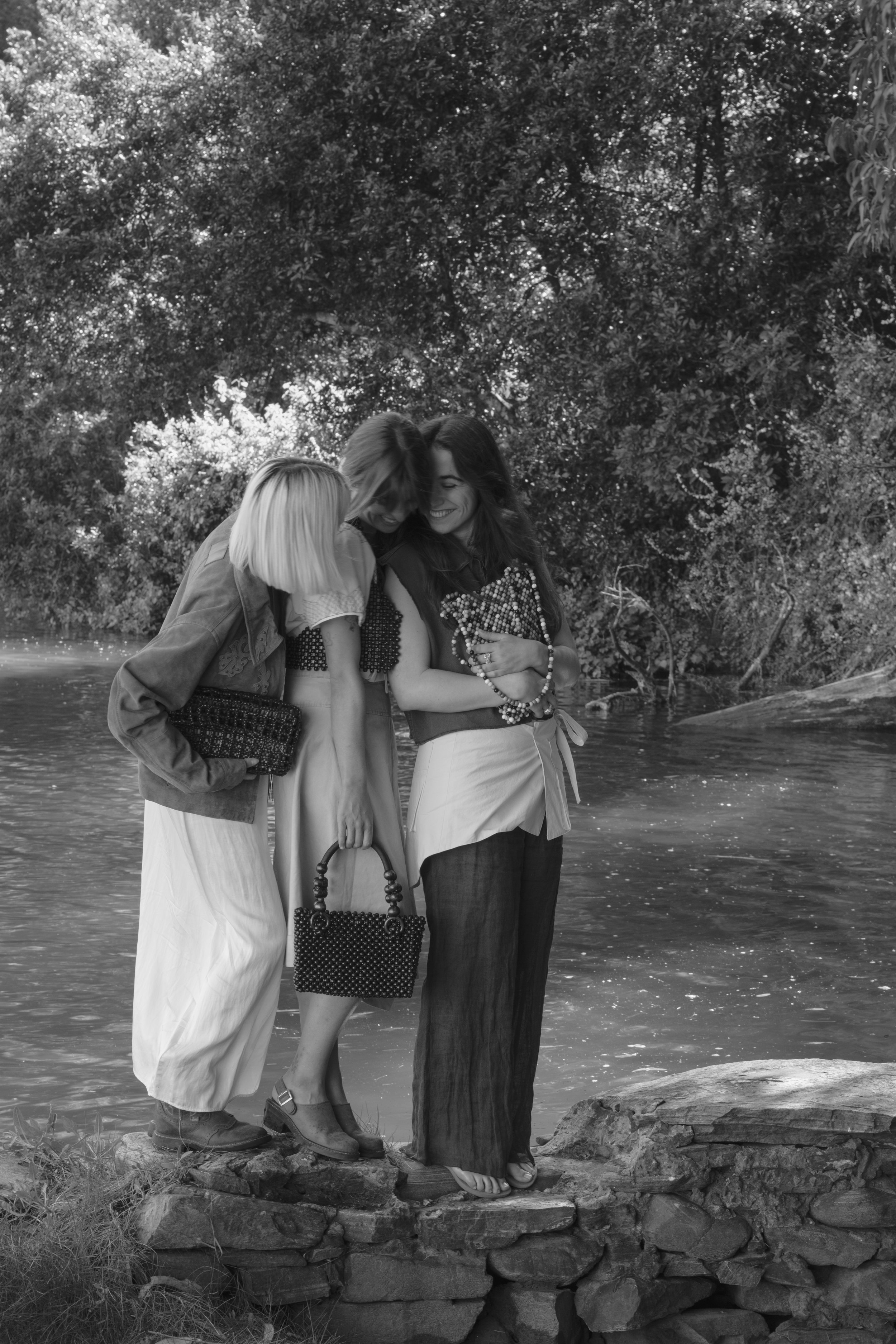 Three women standing on rocks by a river, laughing and embracing each other in a natural, wooded outdoor setting.