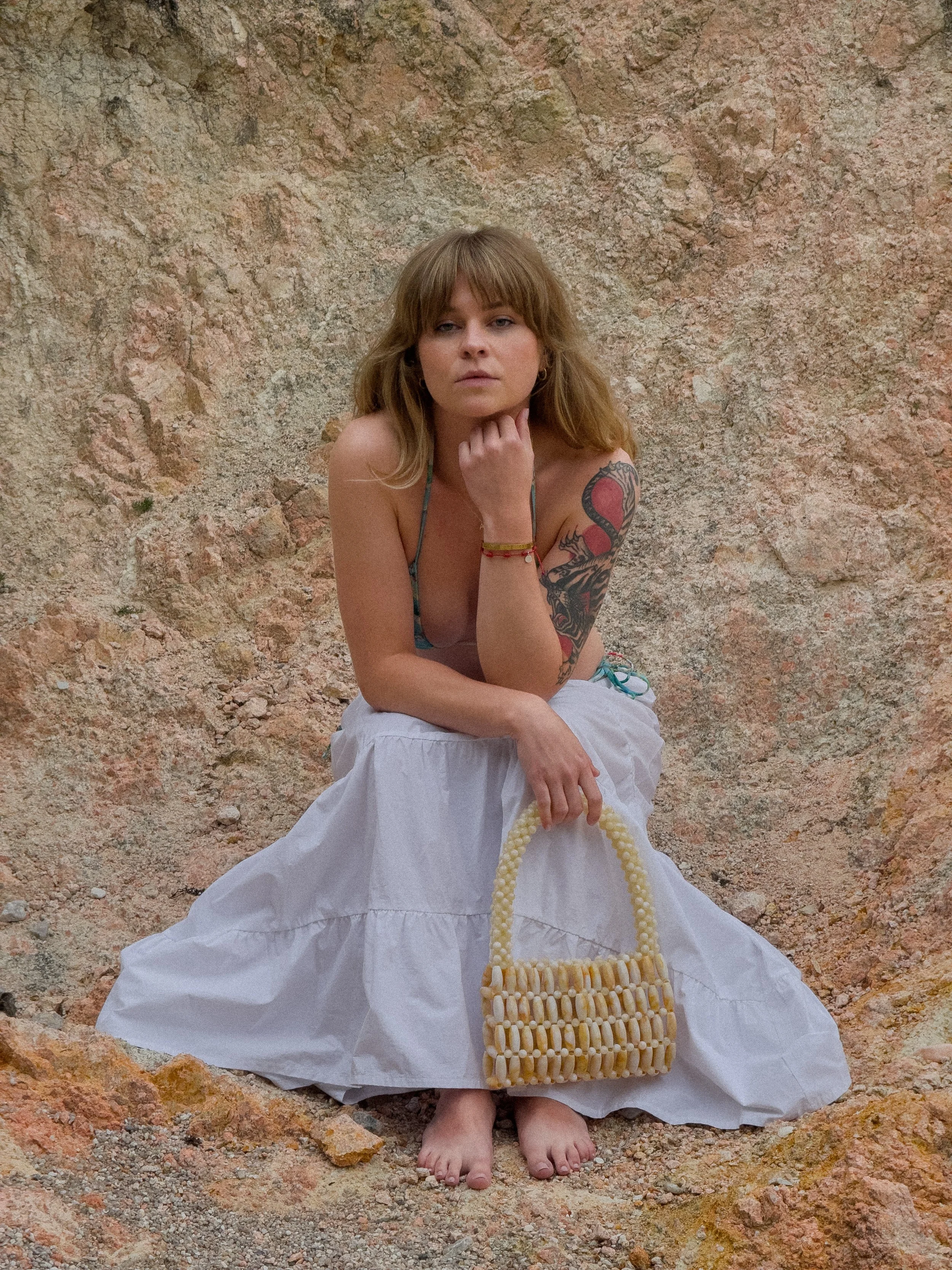 Young woman with tattoos, sitting barefoot on rocky terrain, holding a woven bag, with a neutral expression, against a rocky background.