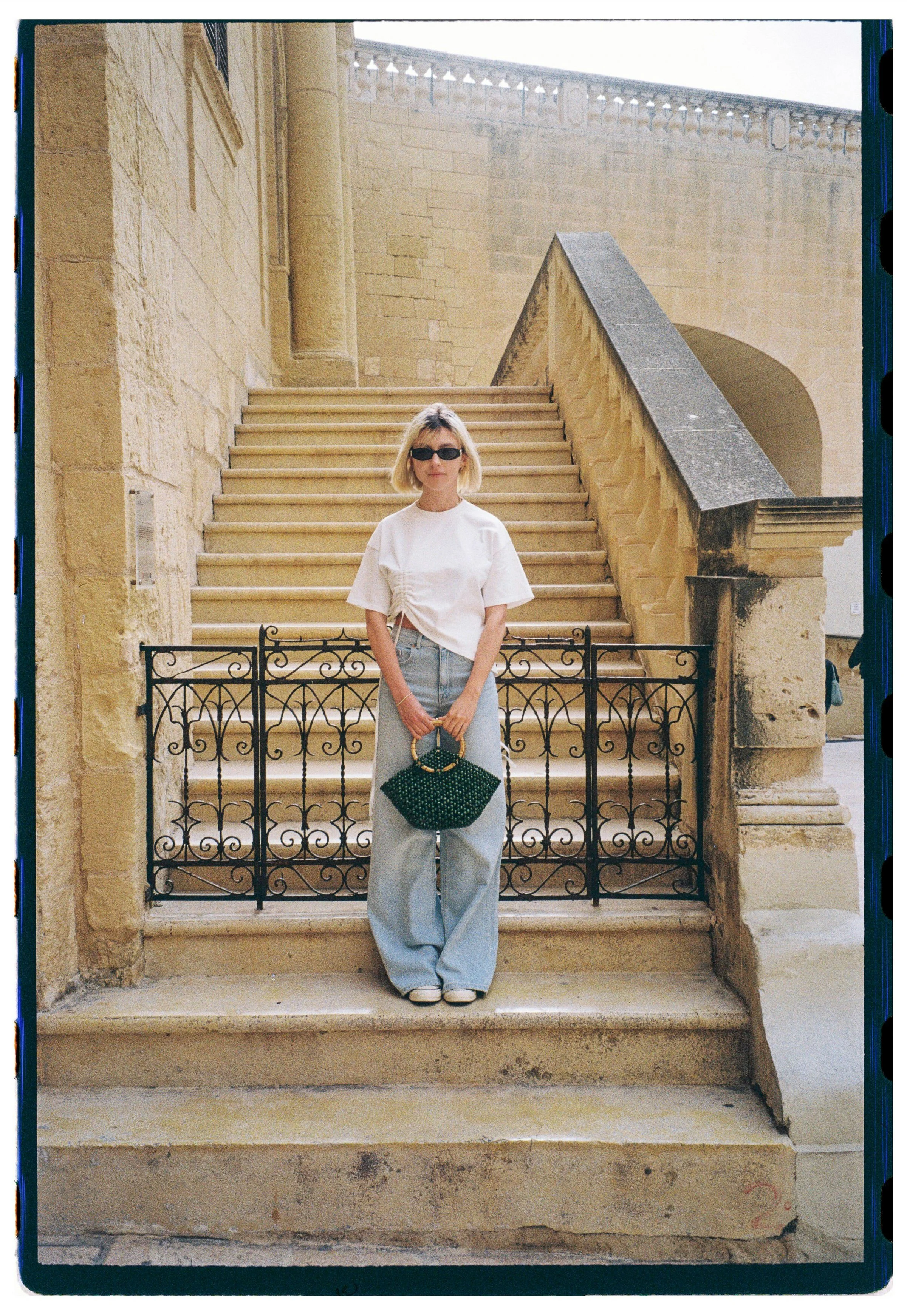 A woman with blond hair wearing sunglasses, a white t-shirt, baggy light blue jeans, and white shoes, stands on stone steps with an ornate black iron railing in front of an old stone building with archways and a staircase.