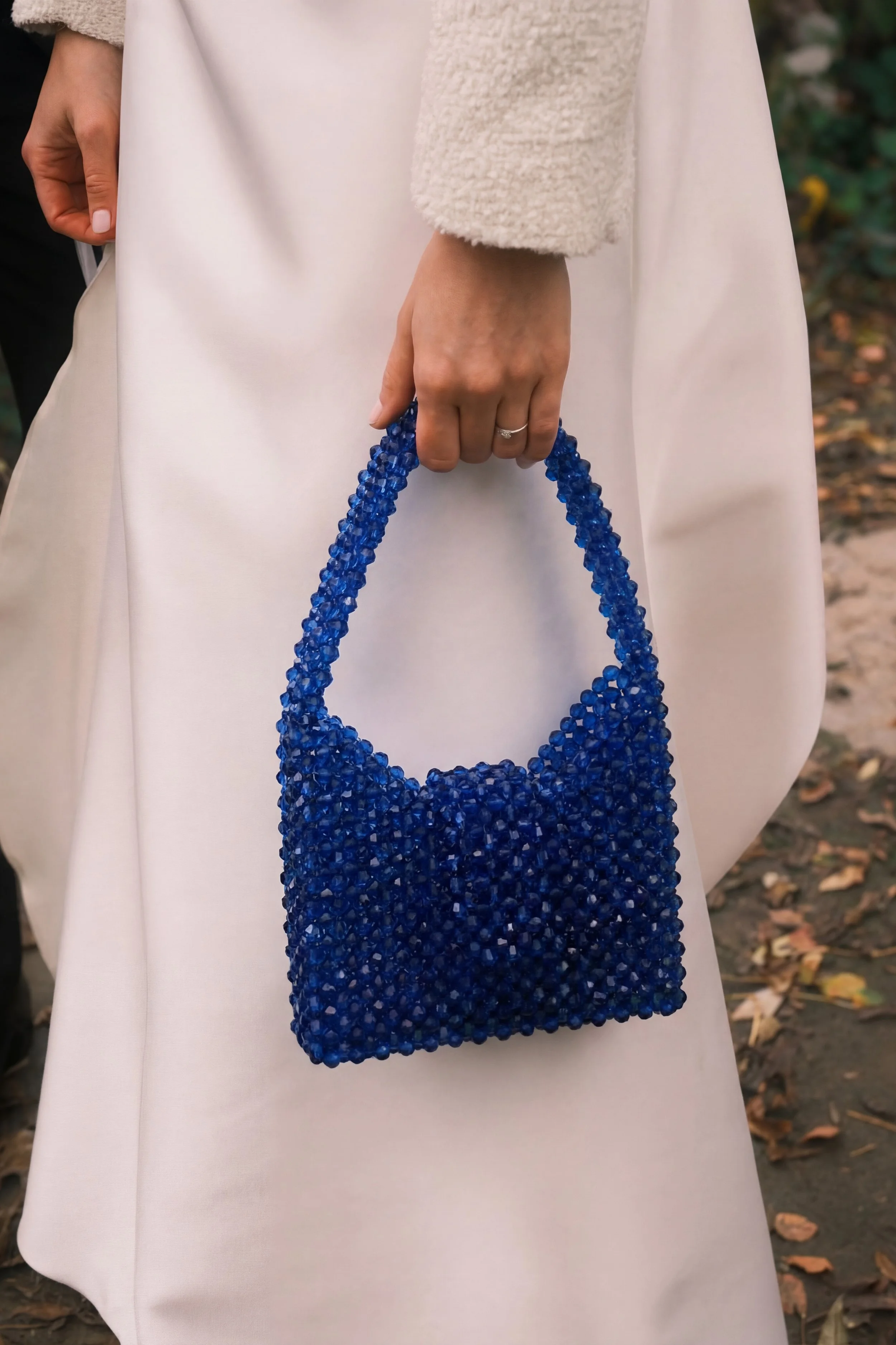 A person holding a small, beaded blue handbag, standing outdoors in a beige coat.