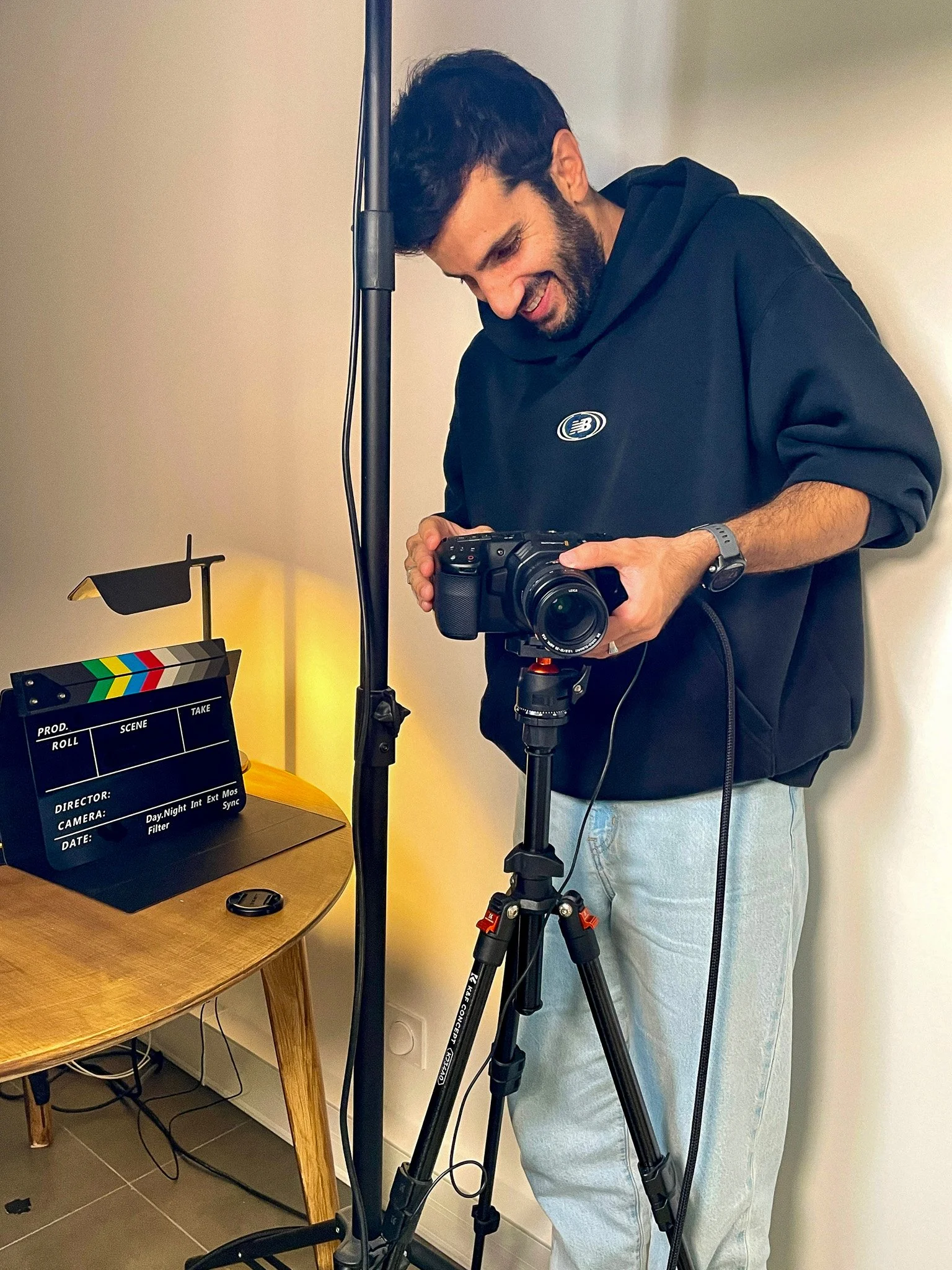 Un homme avec un sweat à capuche noir et un pantalon clair, utilisant un appareil photo monté sur un trépied, dans un intérieur avec un éclairage chaud et une table à côté.