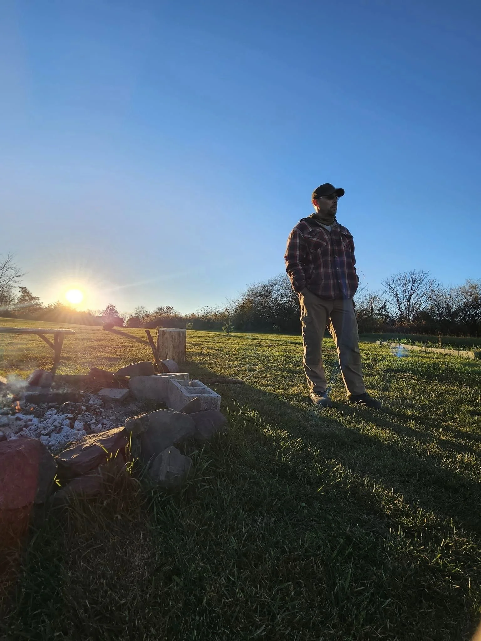 A man standing on grass in a park during sunset, wearing a plaid jacket, glasses, and a cap, with a small fire pit and rocks nearby.