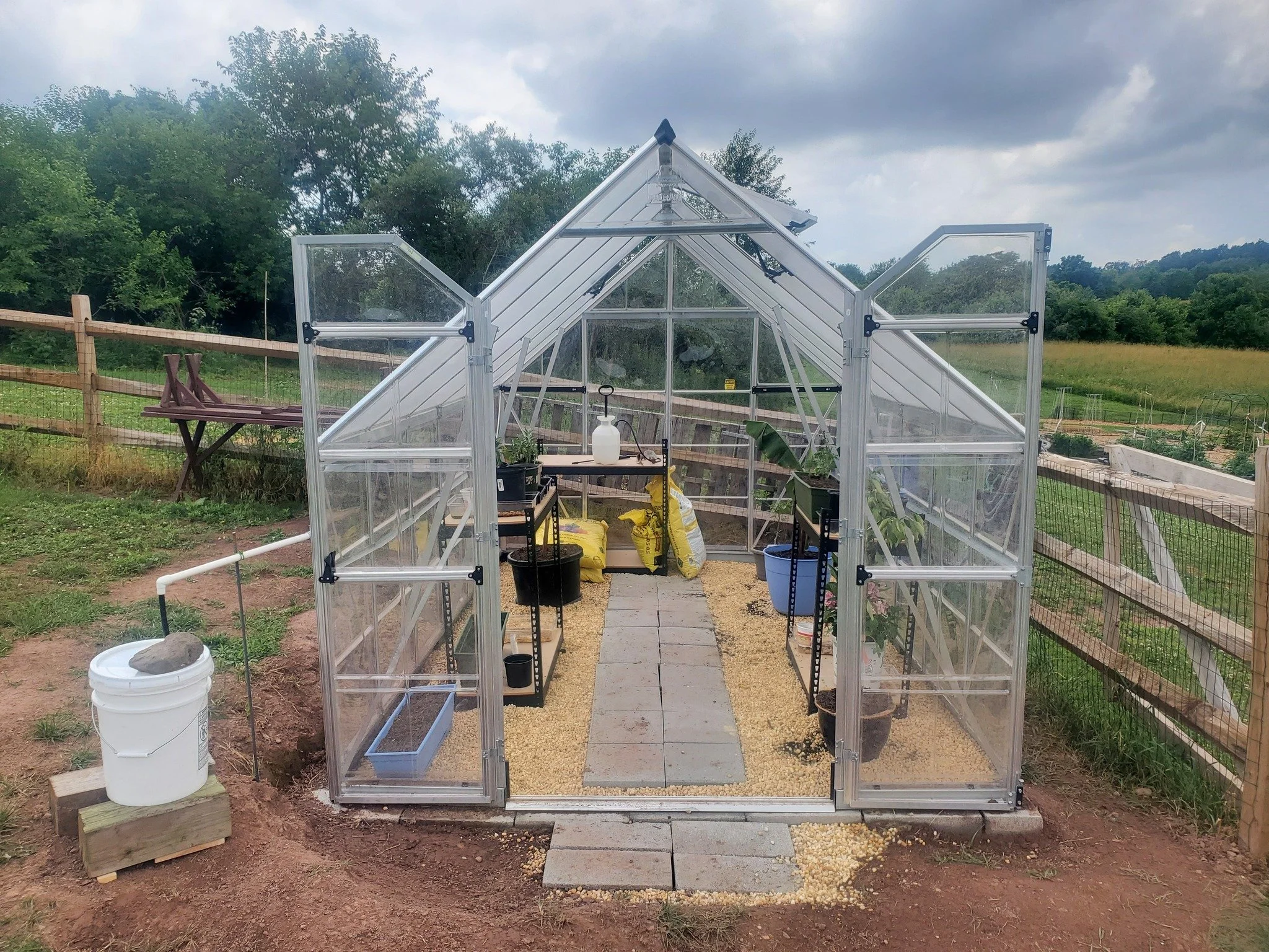 A small greenhouse with transparent walls and roof, filled with potted plants, gardening supplies, and a pathway made of pavers surrounded by gravel, in a rural outdoor setting with a grassy field and trees in the background.