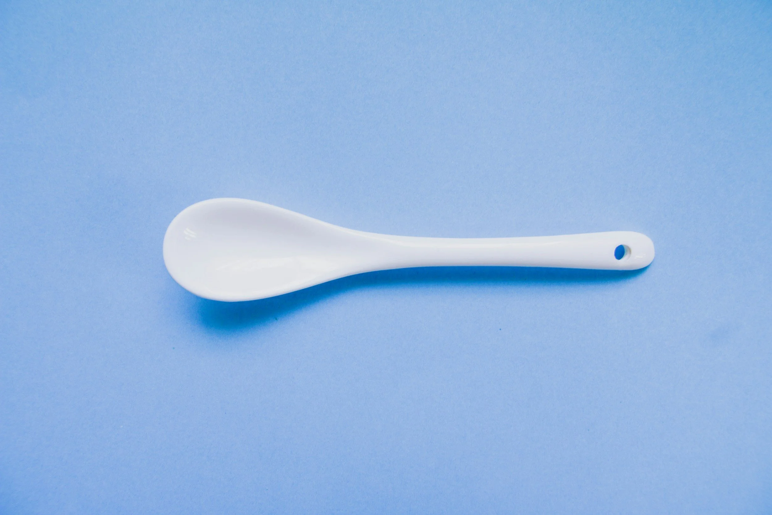 A white ceramic spoon with a blue circle at the handle's hole, placed on a solid light blue background.