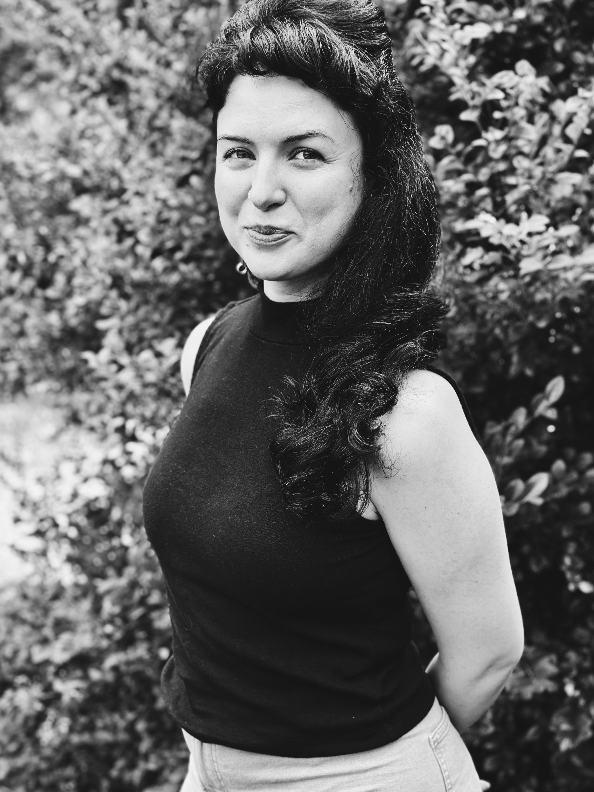 A woman with dark, wavy hair pulled back, wearing a sleeveless top, stands outdoors in front of a leafy background, smiling slightly with her lips closed.
