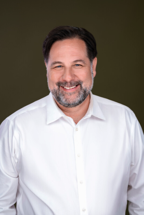 A smiling middle-aged man with dark hair and a beard, wearing a white button-up shirt, standing against an olive green background.
