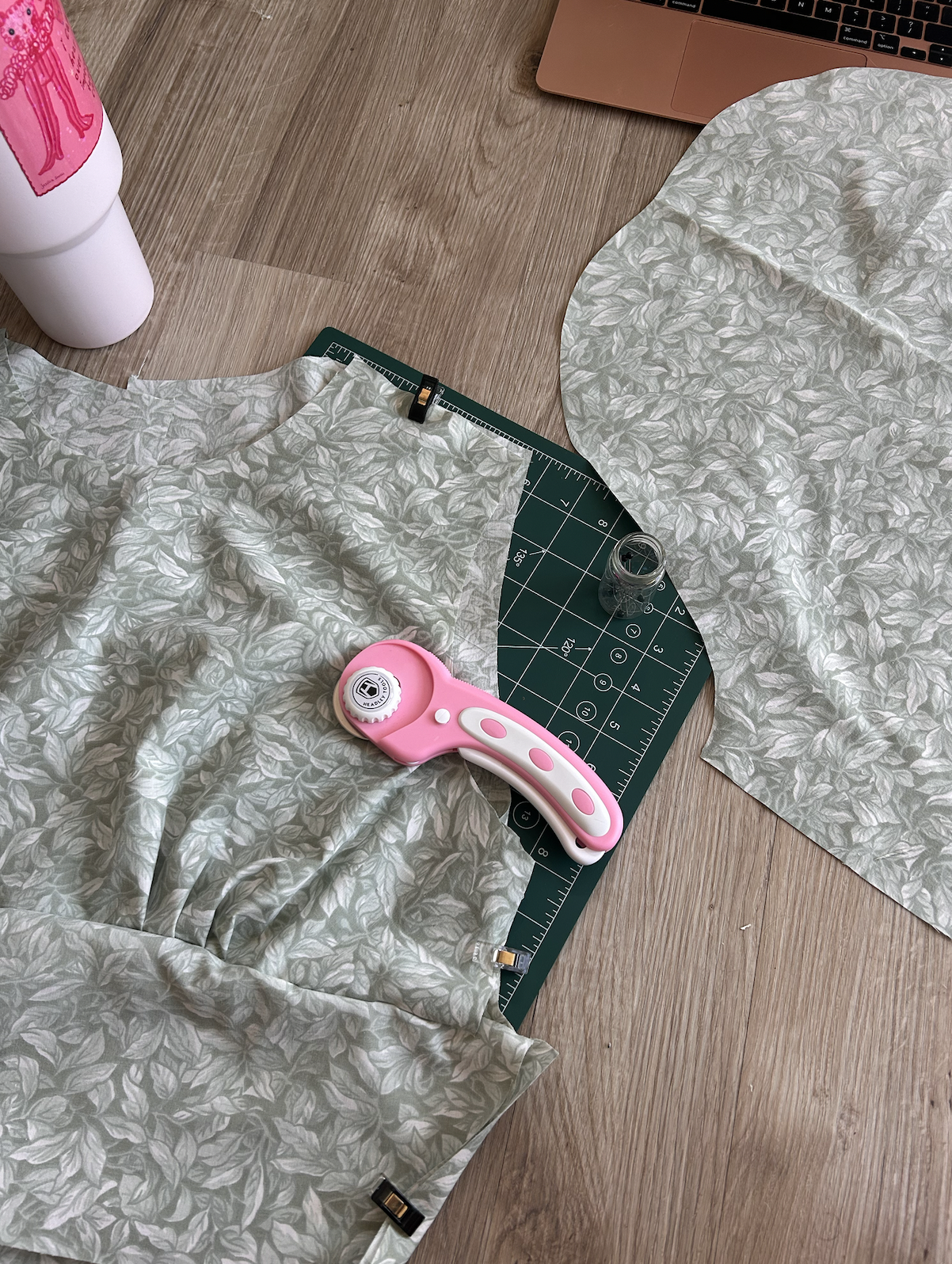 Fabric with a light green leafy pattern, pink rotary cutter, glass jar, cutting mat, laptop, and water bottle on a wooden table.