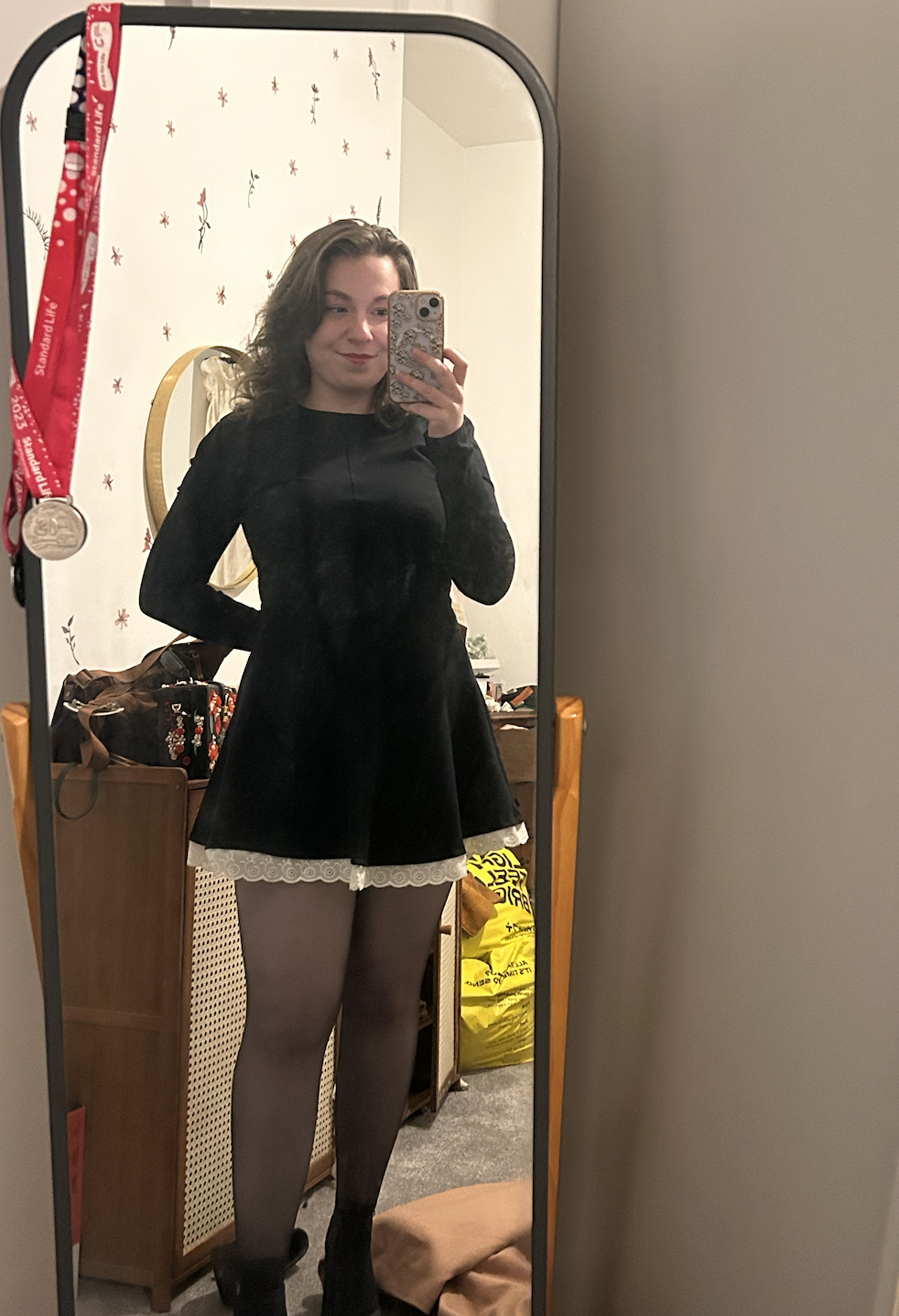 A young woman taking a mirror selfie in her bedroom. She is wearing a black dress with mesh stockings and black shoes. The room has a wooden dresser, a yellow bag, and a wall decorated with small floral patterns.