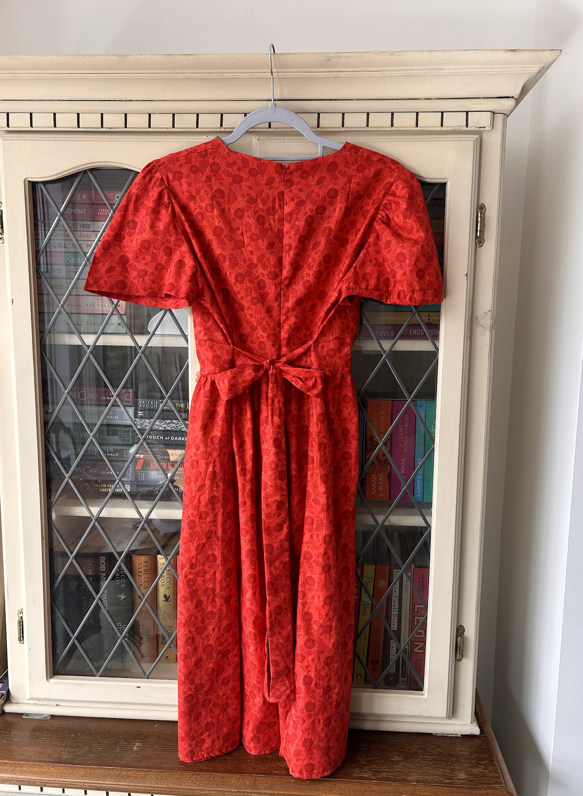 Red dress with puffed sleeves and a bow at the waist, hanging on a hanger in front of a cabinet filled with books.
