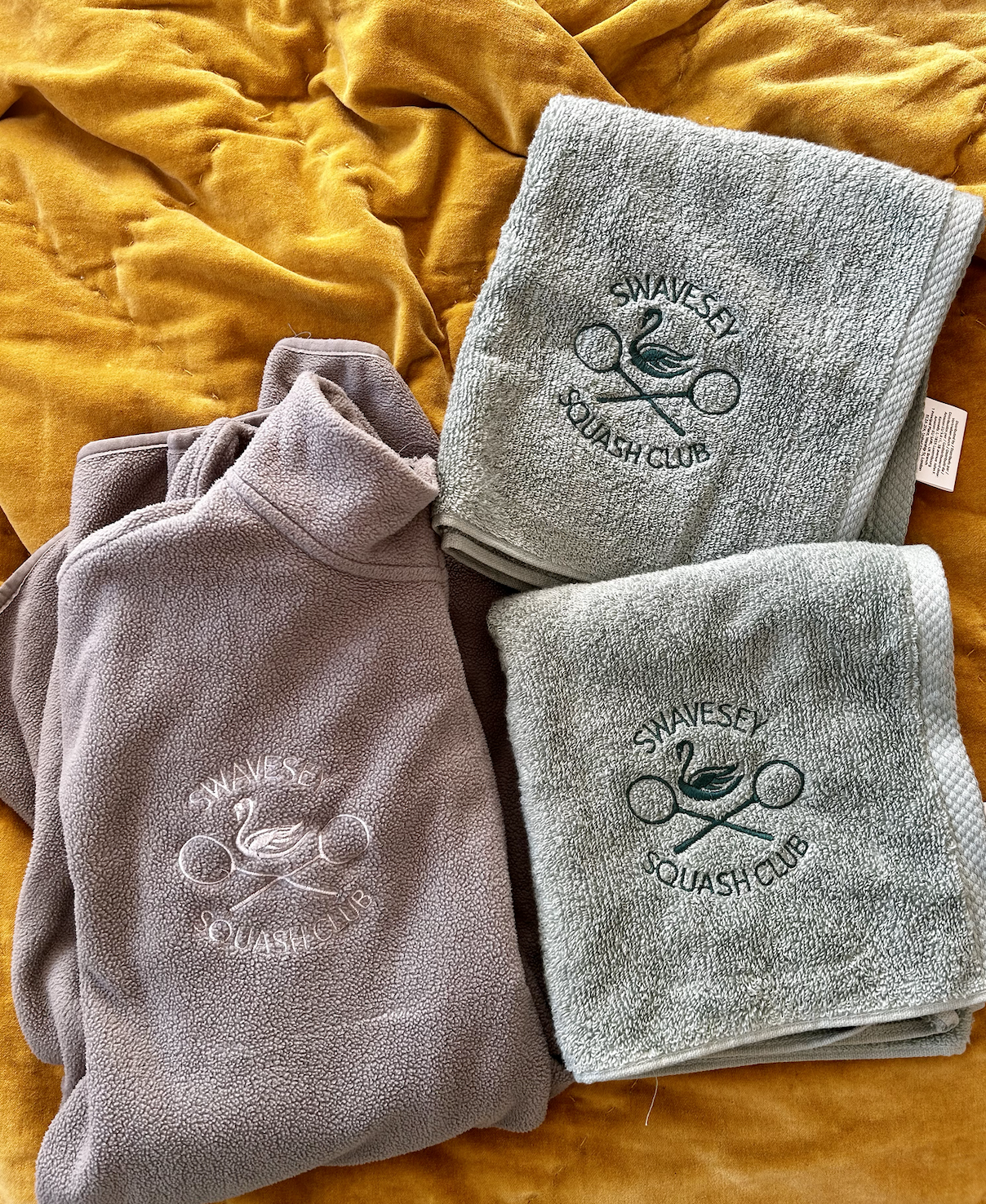Three towels and a fleece jacket with 'Swavesey Squash Club' logo, all placed on a yellow fabric background.