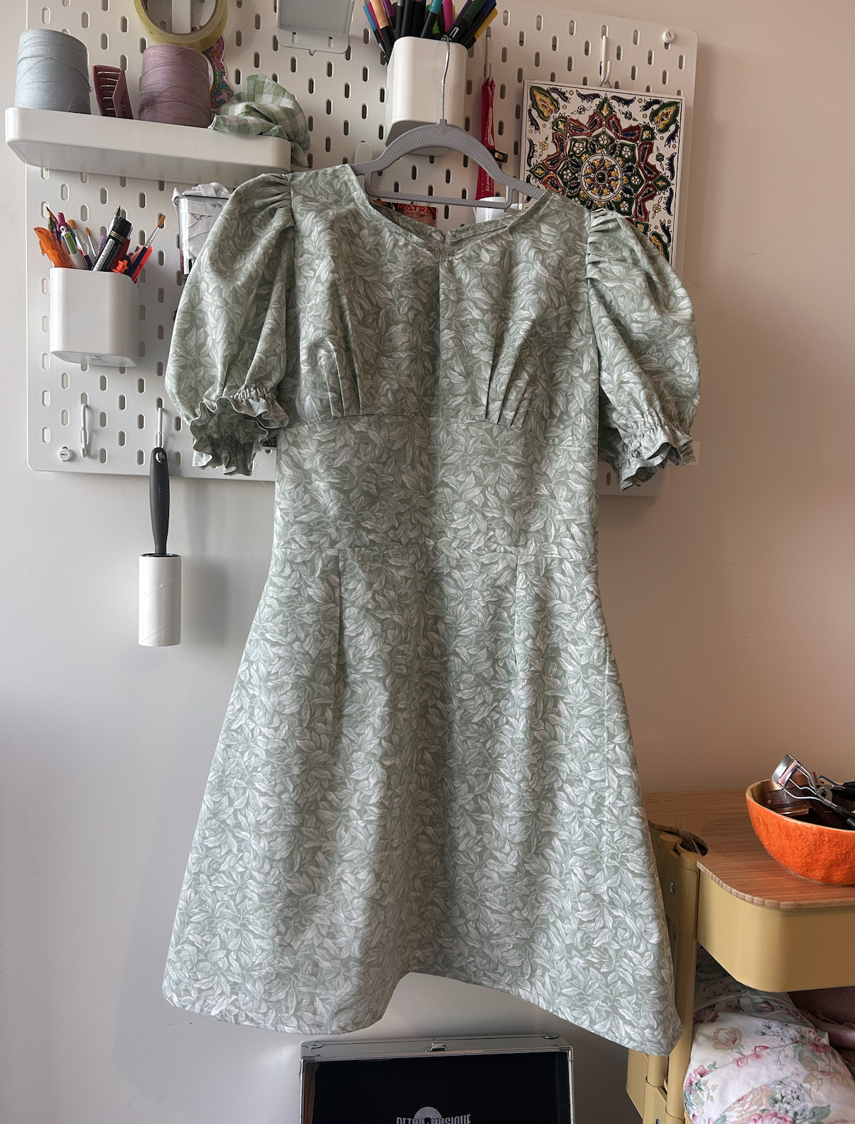 A light-colored dress with a leaf pattern hanging on a white pegboard, surrounded by art supplies and tools.