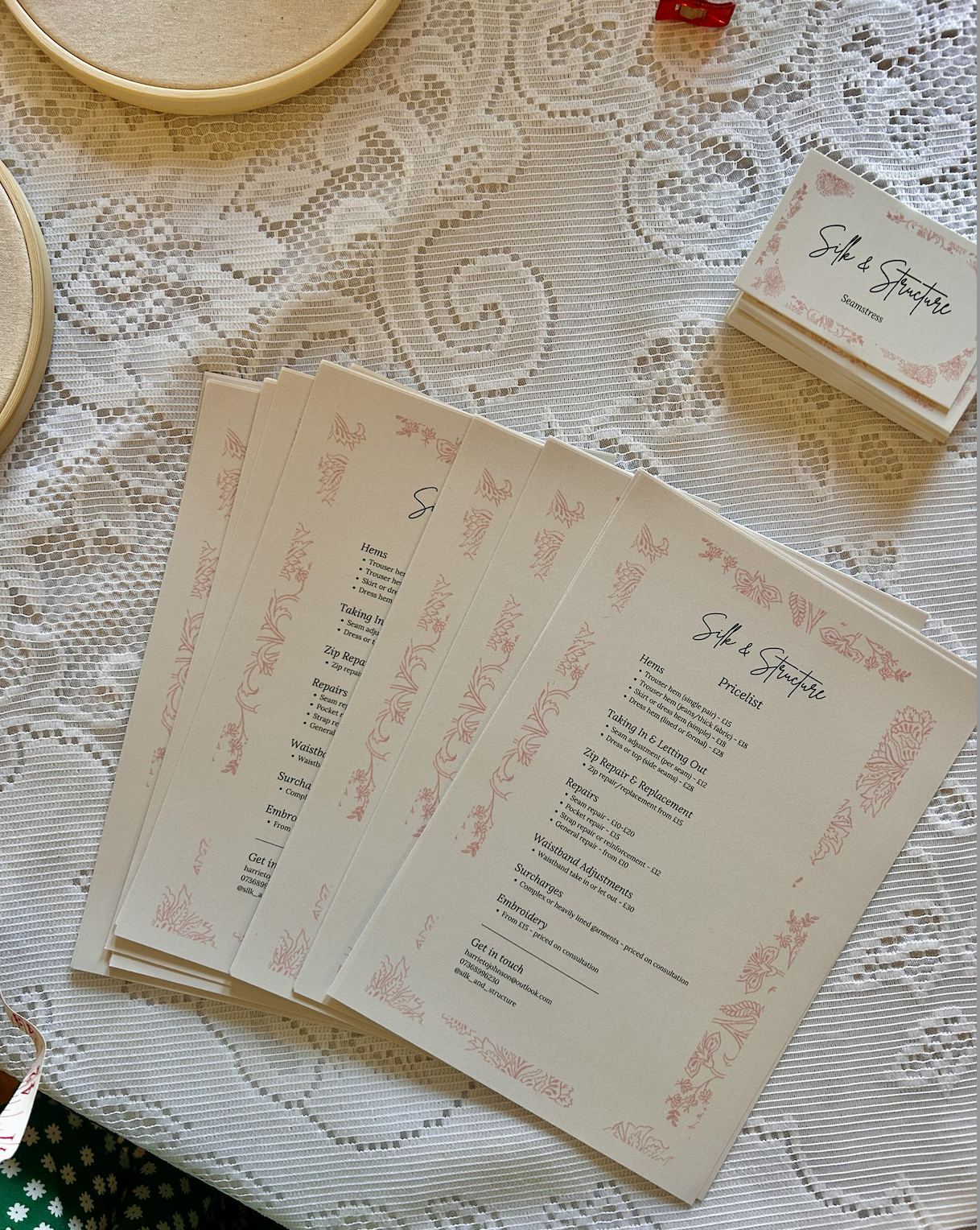 Set of printed menu cards titled 'Silk & Structure' on a lacy fabric tablecloth, with stacks of cards and organizational containers nearby.