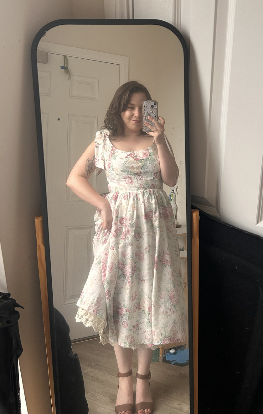 Woman in floral dress taking a mirror selfie indoors, with a closed door and wooden floors visible.