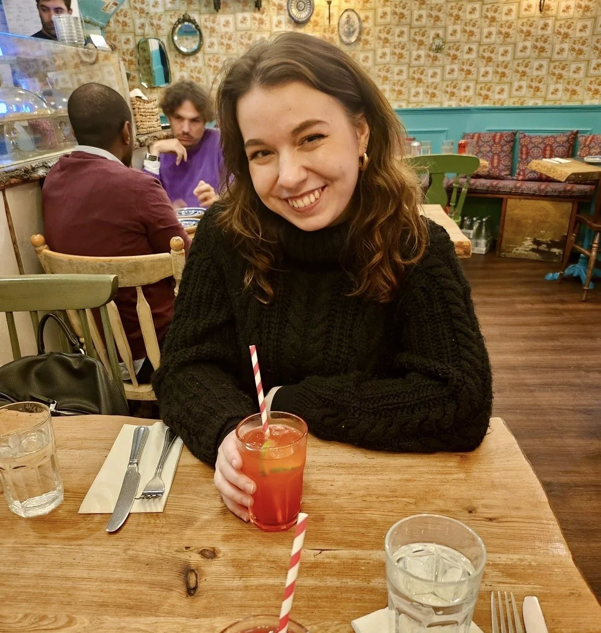 A woman with shoulder-length brown hair smiling and sitting at a wooden table in a restaurant, holding a glass of red-colored drink with a straw, wearing a black knit sweater.