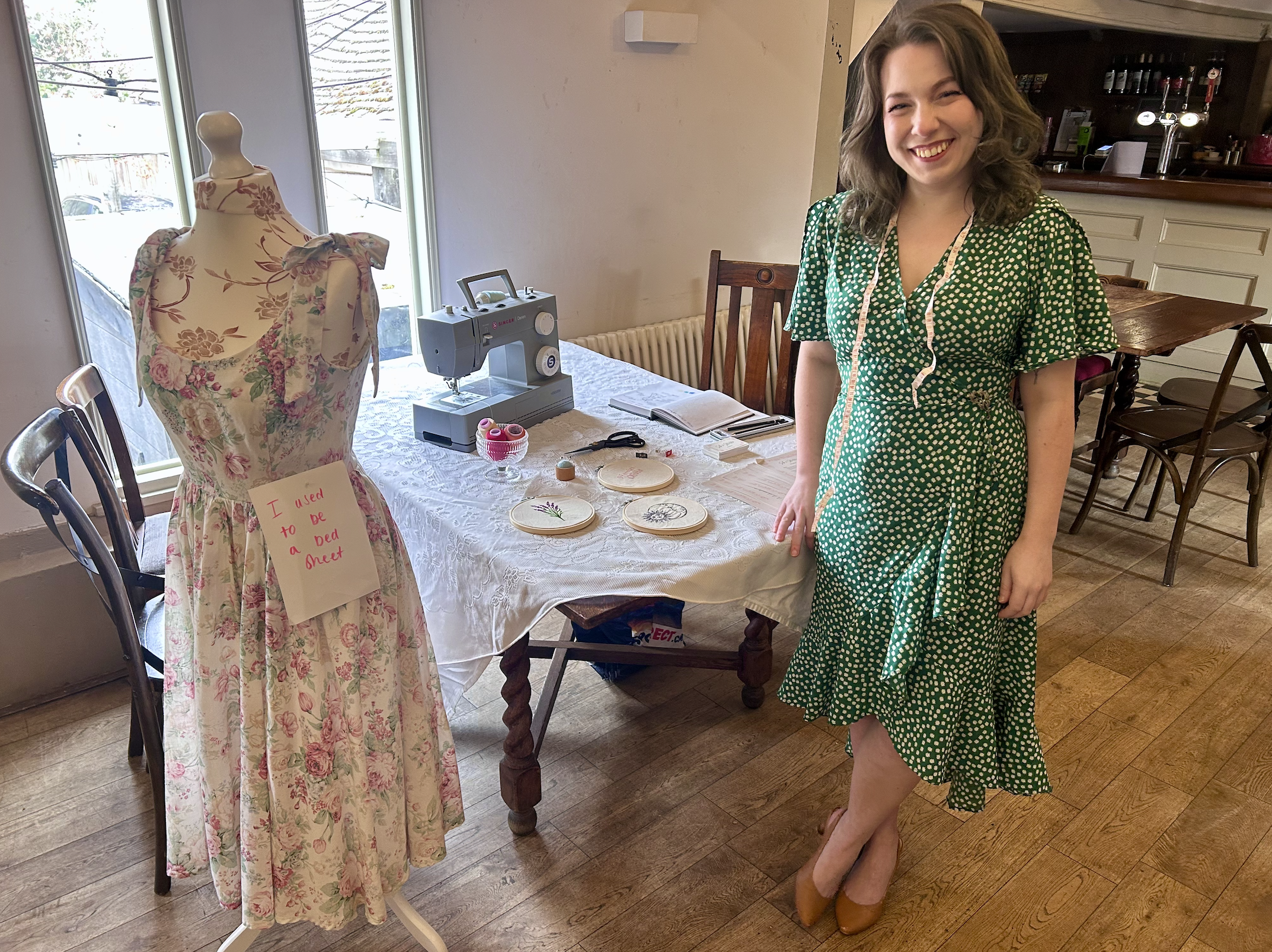 Woman in a green polka dot dress smiling standing next to a mannequin wearing a floral dress with a handwritten sign that says, "I used to be a Bed Meet." The table behind them has embroidery hoops, fabric, scissors, and a sewing machine, set in a cozy room with wooden flooring and large windows.