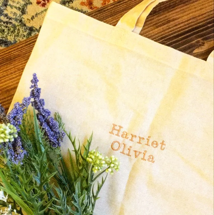 A cream-colored tote bag with a yellowish hue displaying embroidered text reading "Harriet Olivia." Next to the bag, there are purple and yellow flowers with green stems and leaves, set on a wooden surface.