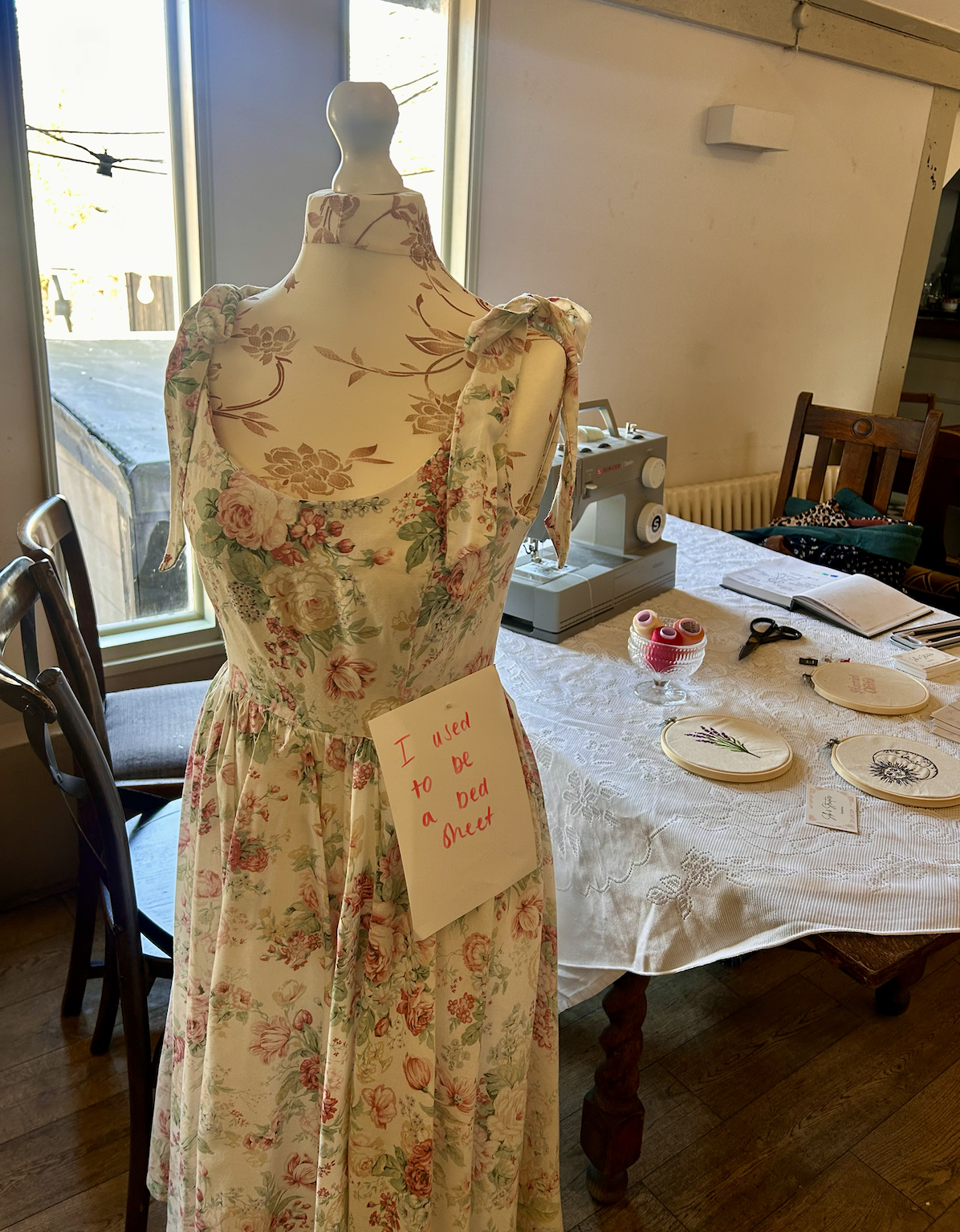 A mannequin wearing a floral dress with a note that says 'I used to be a dress sheet'. The setting is inside a room with a table covered in embroidery supplies, sewing machines, and notebooks, near a window.