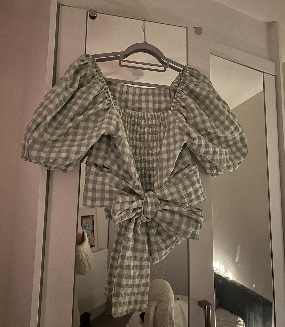 A short puff sleeve, plaid shirt with a tied knot at the front, hanging on a hanger in front of a mirror.