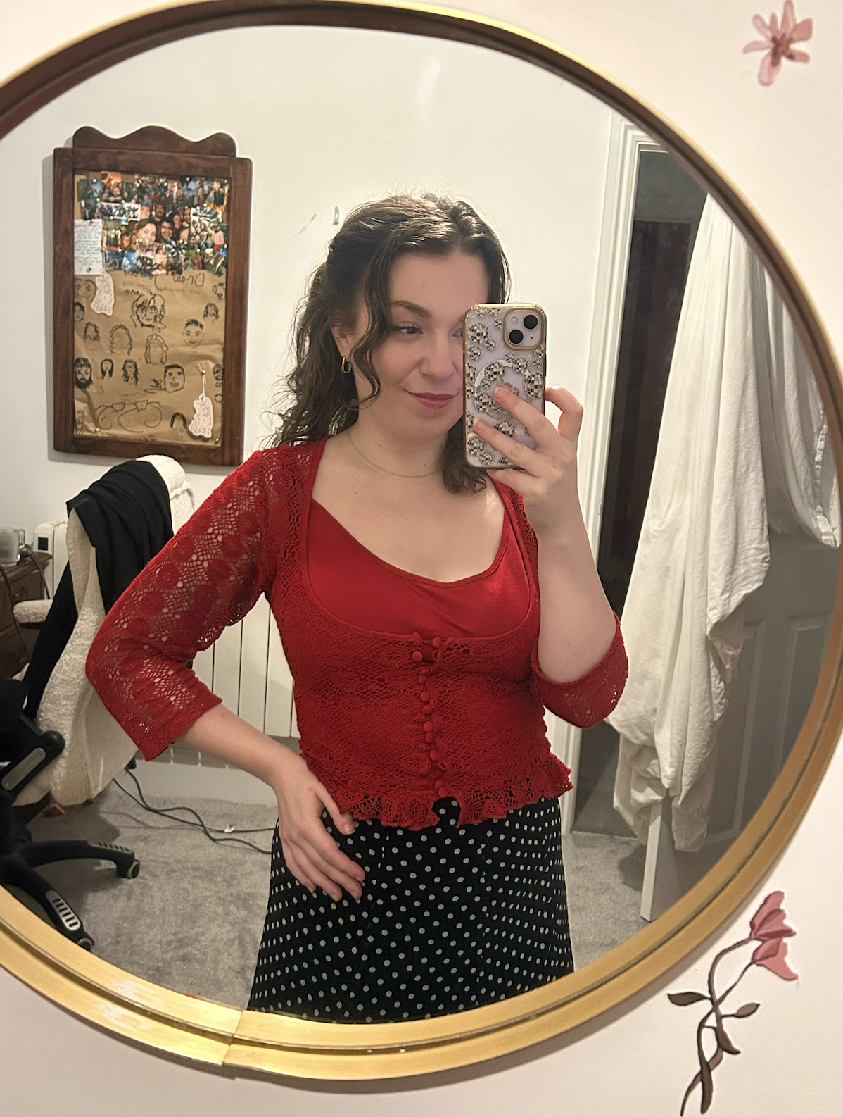 A woman taking a mirror selfie in a bedroom. She is wearing a red lace top and a black skirt with white polka dots, and has curly brown hair.