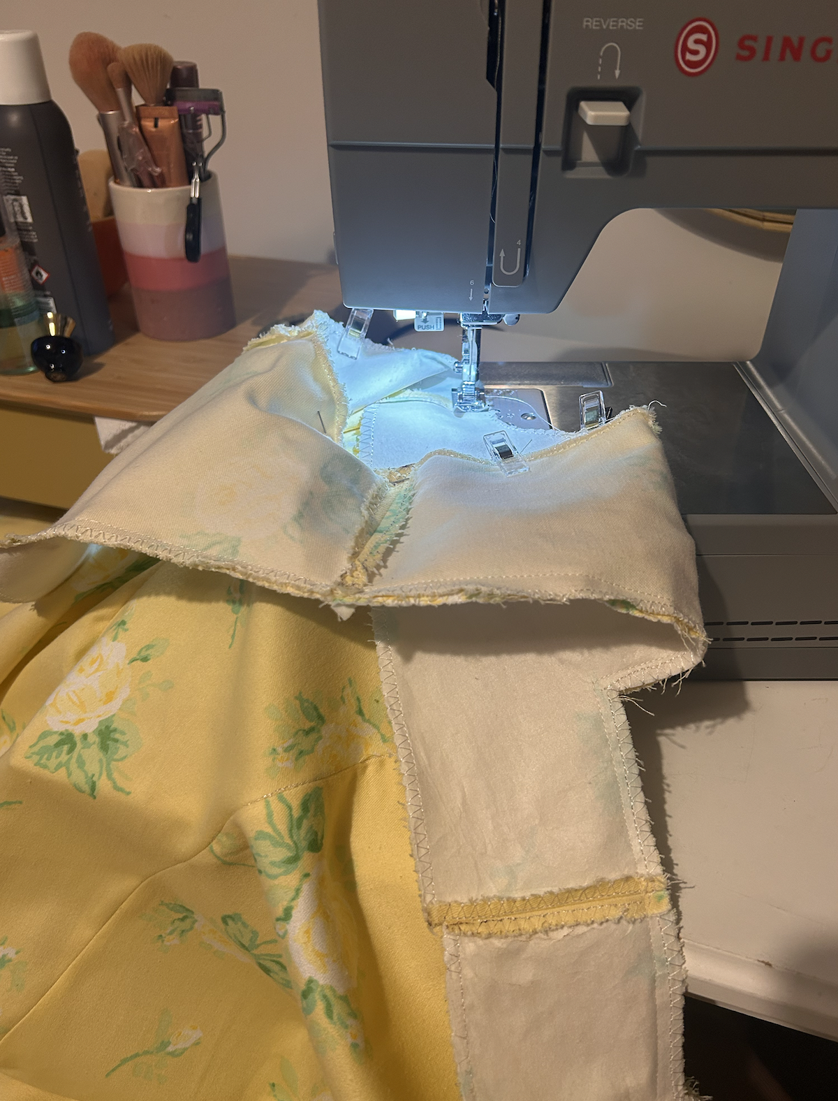 A sewing machine stitching yellow fabric with floral green and white pattern, with fabric pieces underneath. A side table with various sewing tools and supplies is visible in the background.