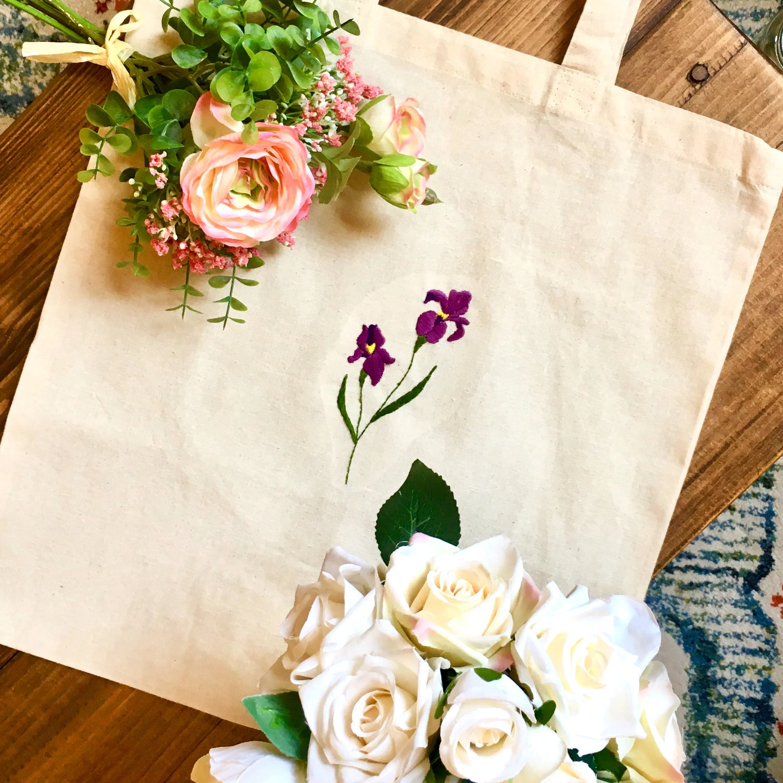 Floral arrangement with pink roses and green foliage, embroidered purple irises on a beige fabric tote bag, and white roses at the bottom.