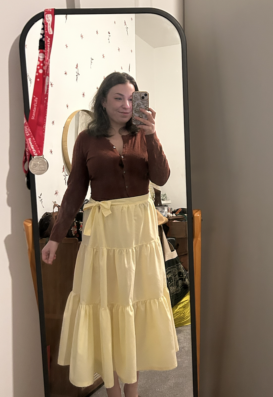 A woman taking a mirror selfie, wearing a brown long-sleeve top and a yellow tiered maxi skirt with a bow tied at the waist.