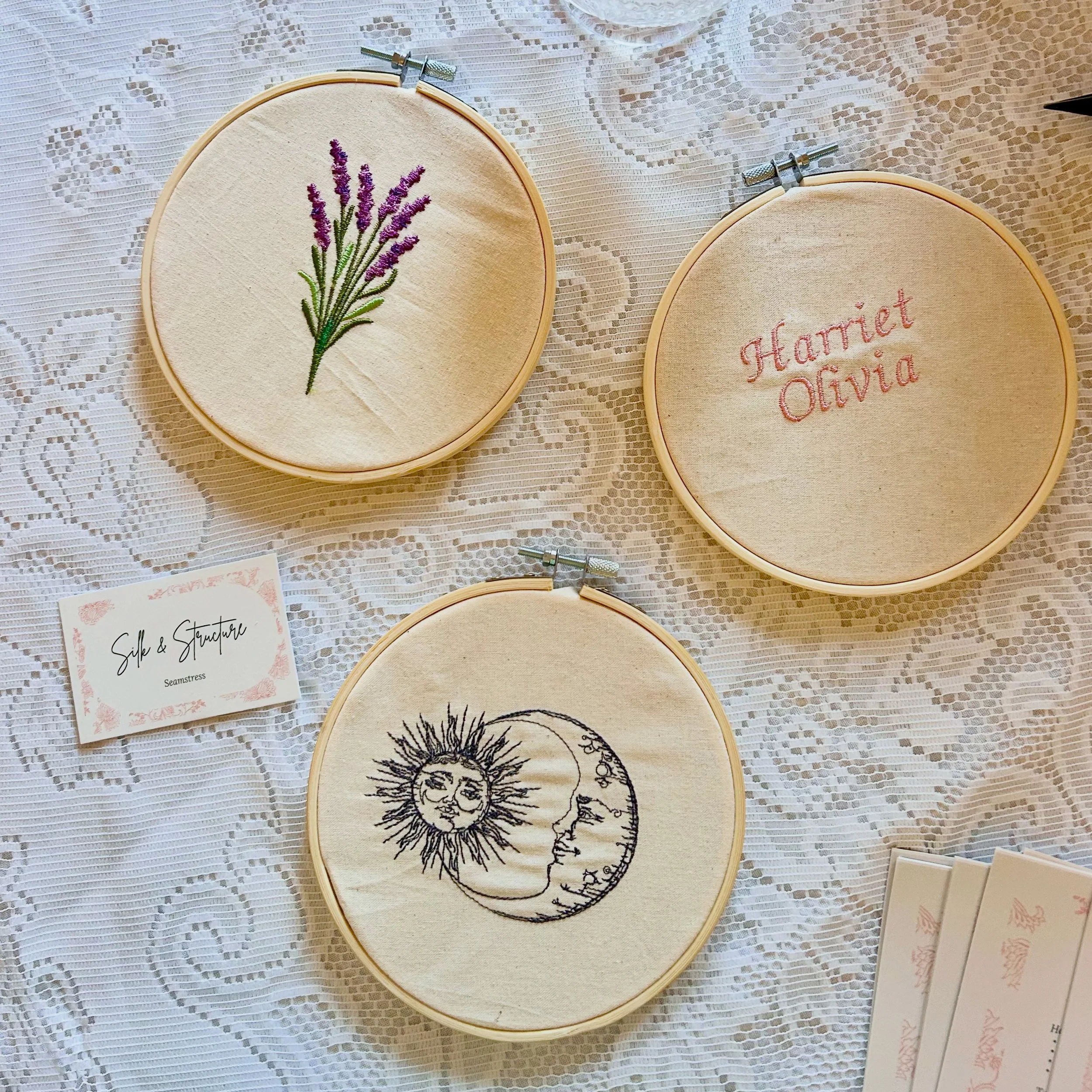Three embroidery hoops with different designs displayed on a lace tablecloth. The top left hoop has a lavender flower, the top right hoop has the name 'Harriet Olivia' stitched in pink, and the bottom hoop has a drawing of a sun and moon face with st