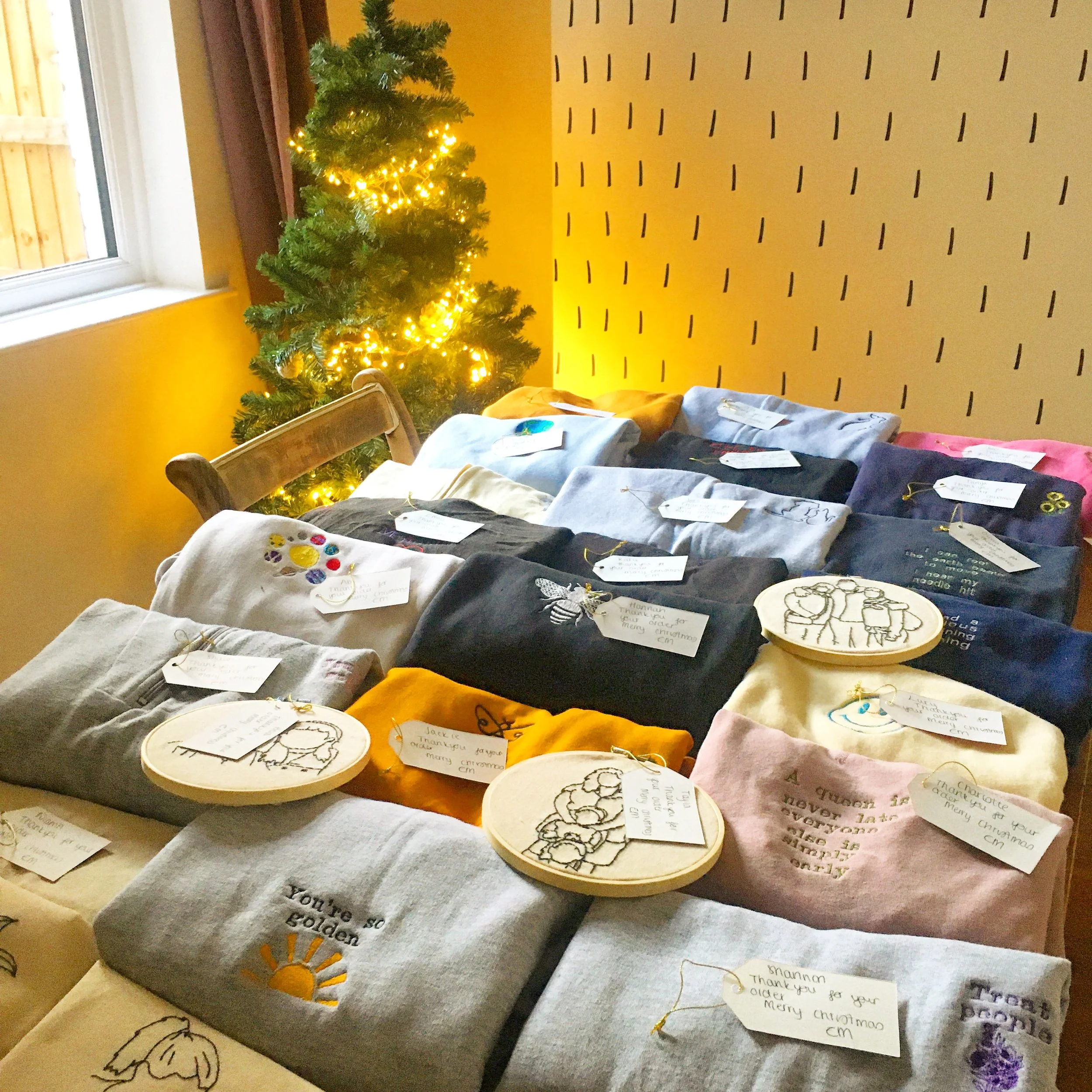 Display of embroidered and decorated holiday shirts and accessories on a table, with a decorated Christmas tree with yellow lights in the background.