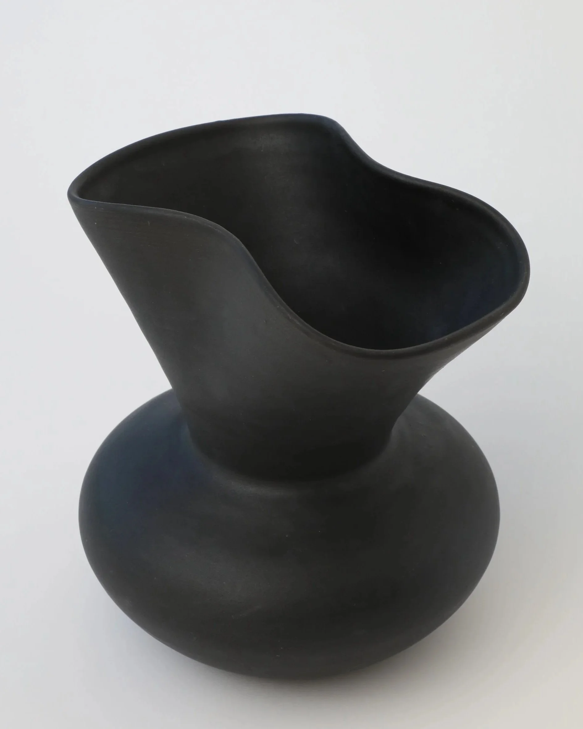 A black, matte-finished ceramic vase with a rounded base and a unique, asymmetrical opening at the top, set against a plain white background.