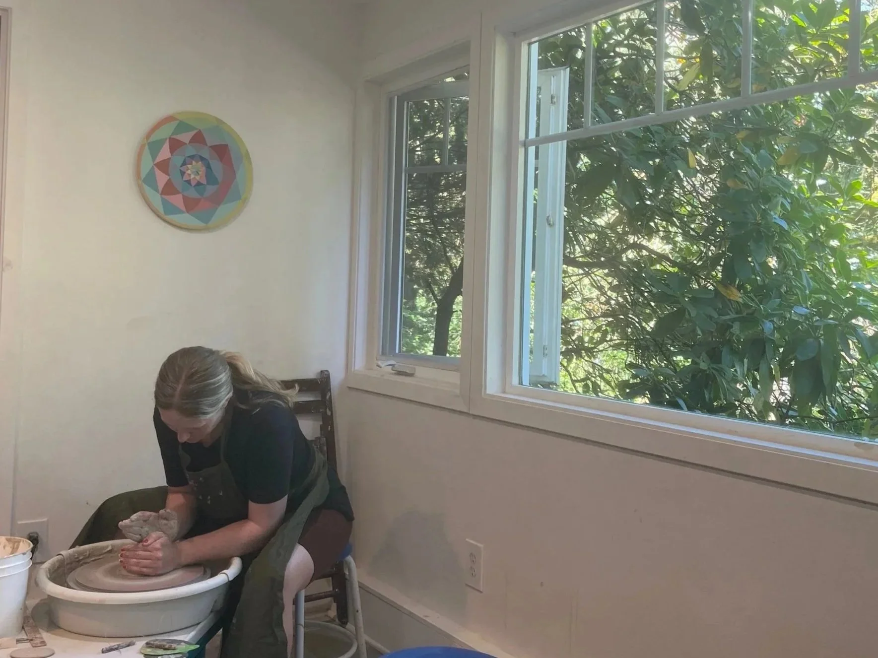 Woman working on pottery at a wheel in a room with open windows and a view of green trees outside, a colorful geometric art piece hangs on the wall.