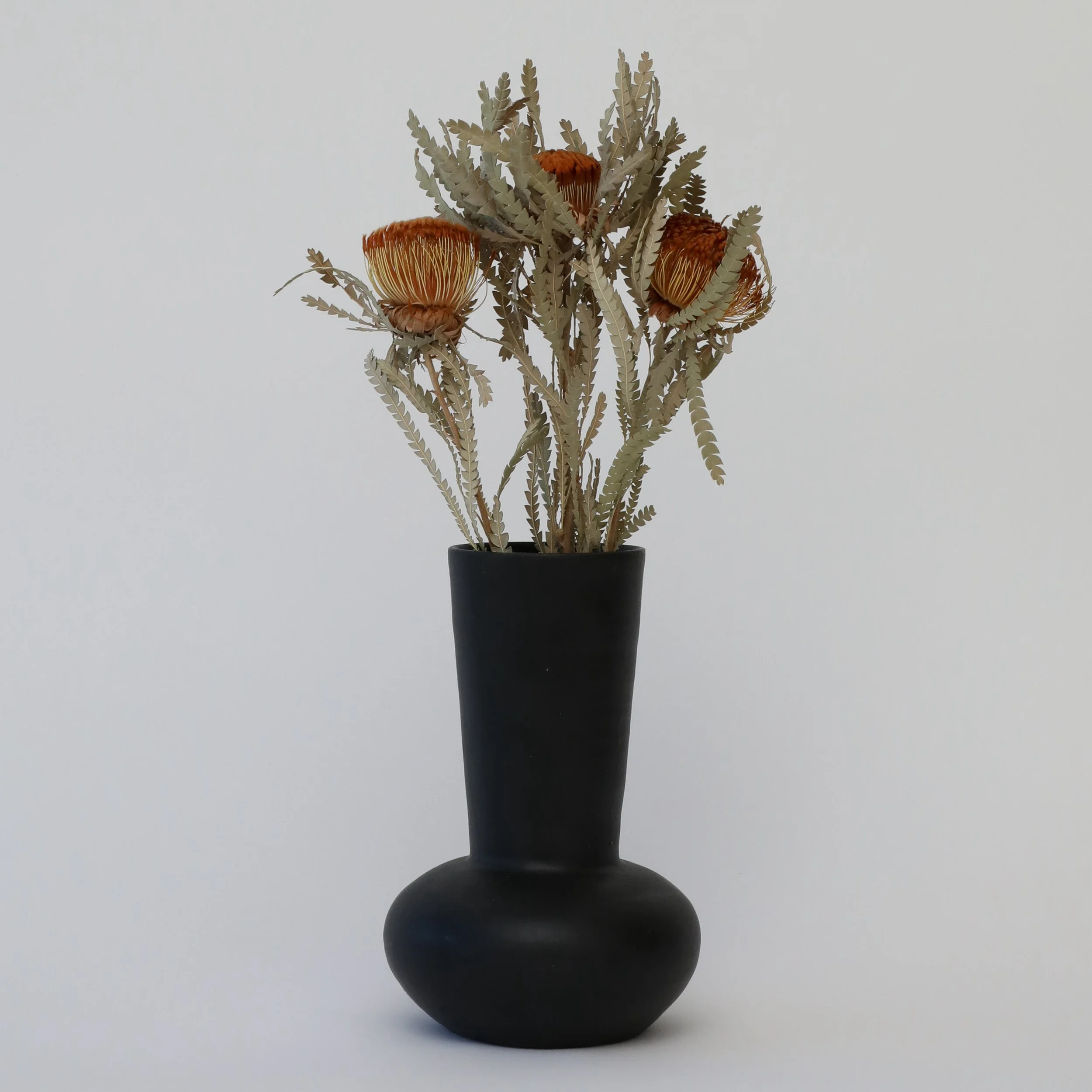 A decorative tall black vase containing dried flowers and foliage, including orange protea flowers and green fern-like leaves.