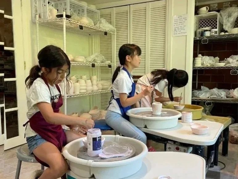 Three young women are seated at pottery wheels, shaping clay in a pottery studio. Shelves in the background hold various ceramic items and supplies.