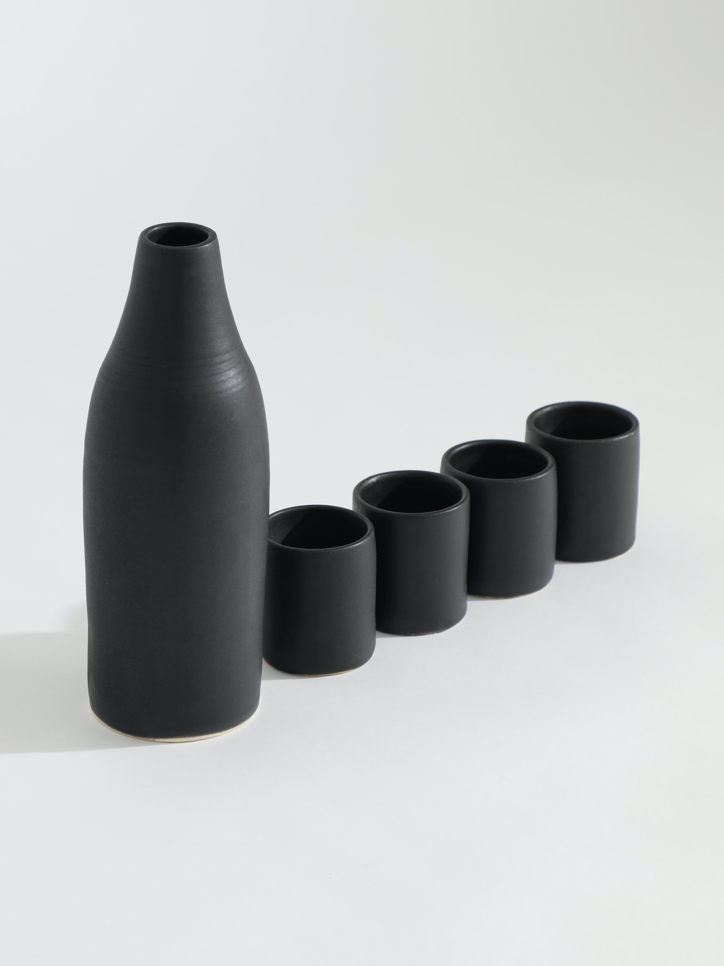 Set of six black ceramic vases arranged in a row on a white surface against a light background, with one taller vase on the left and five shorter vases to the right.