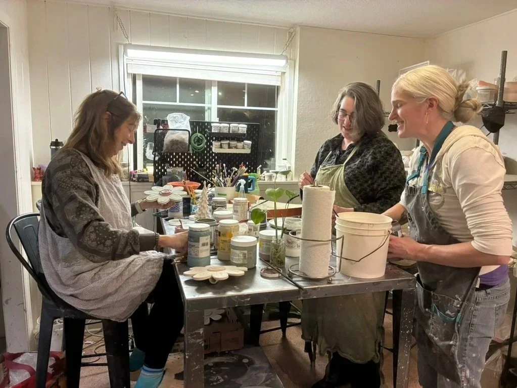 Three women and one girl working together on a craft project at a cluttered table inside a room with a window. The table has various art supplies, paint jars, paper towels, and small ceramic items.