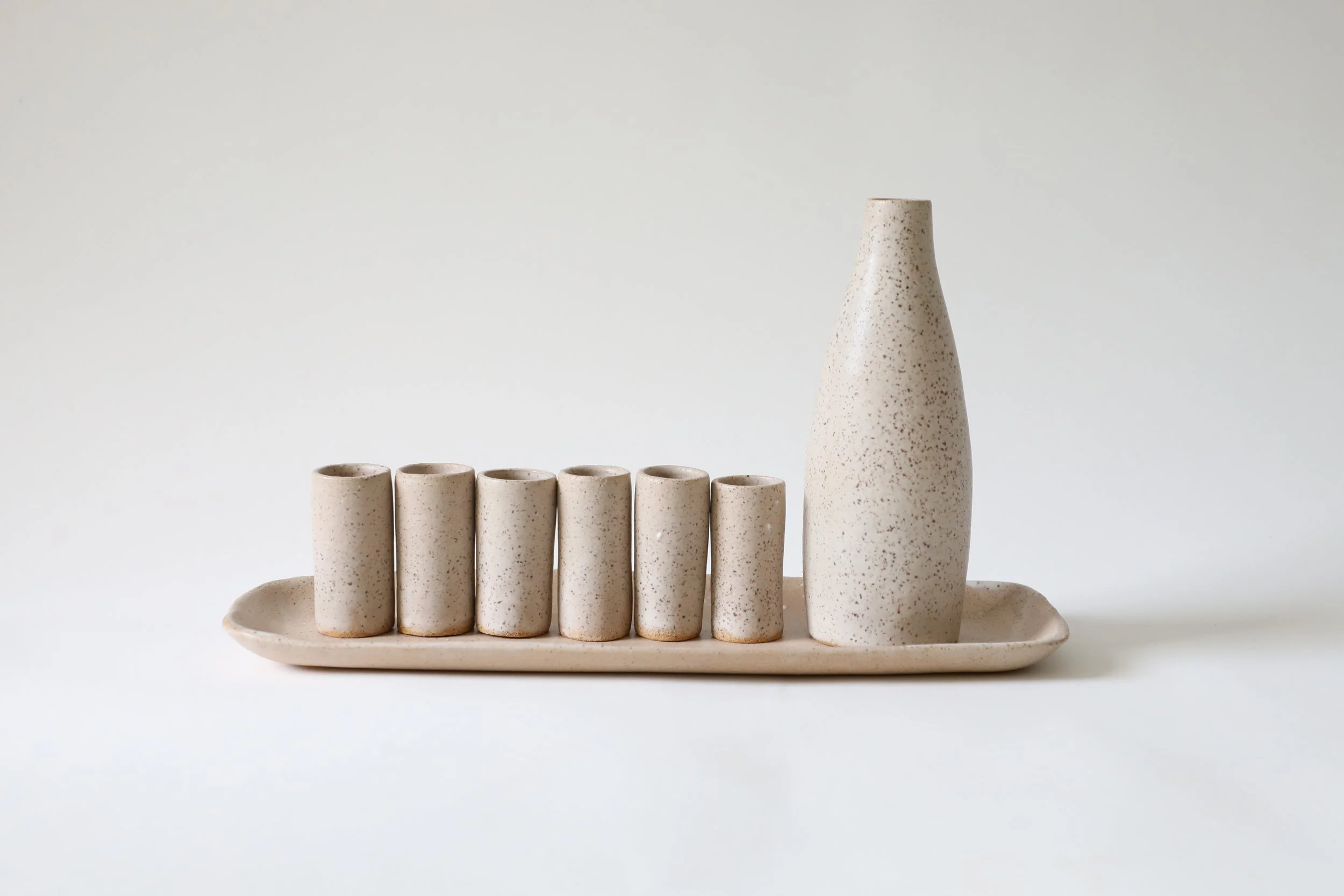 Set of six small beige speckled cups on a matching rectangular tray, and a tall matching vase, all with a minimalist aesthetic.