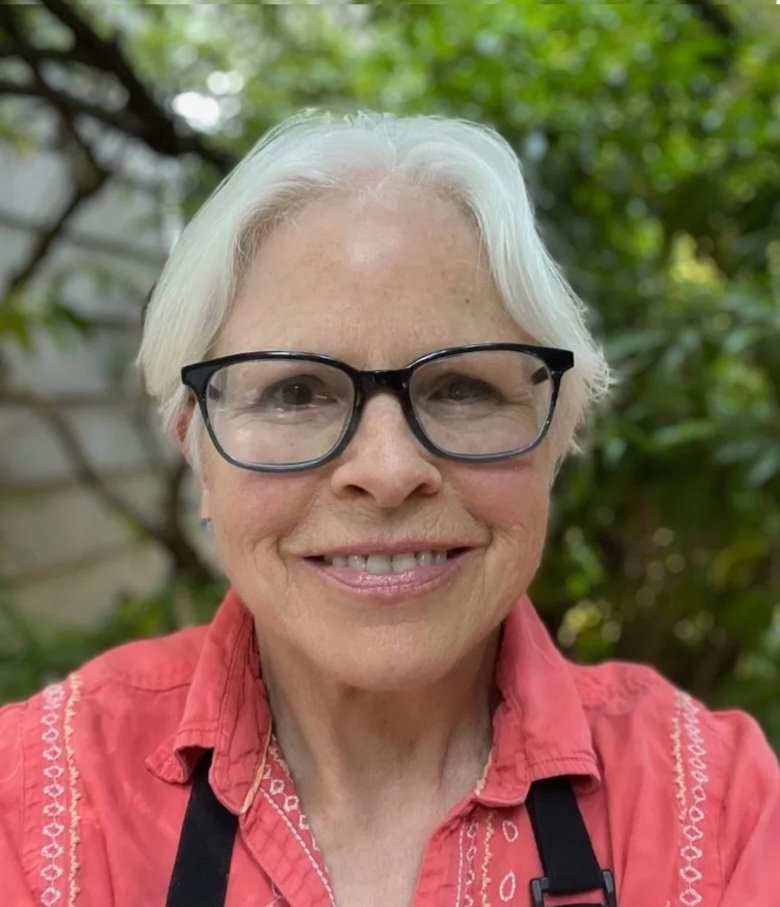 A woman with short white hair, glasses, and a red shirt smiling outdoors, with green foliage in the background.