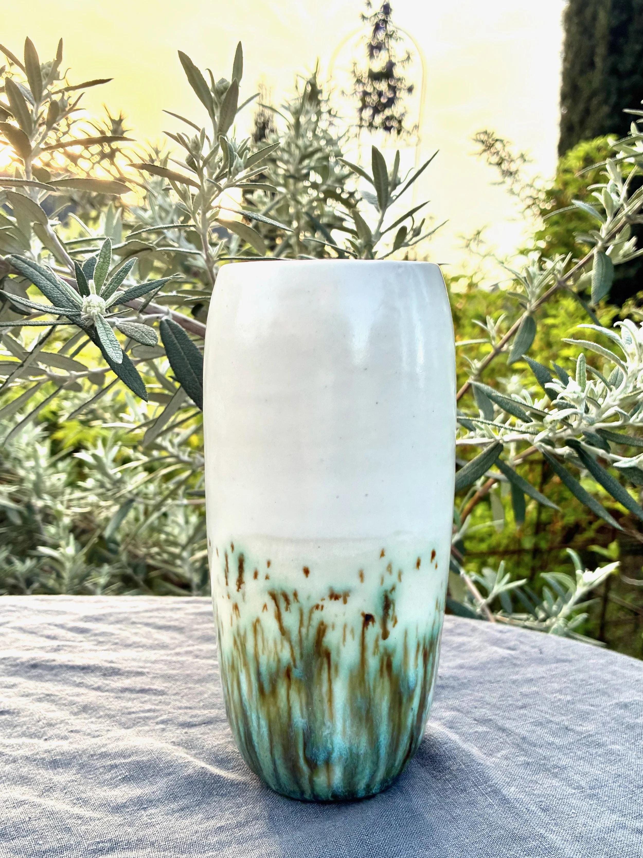 A ceramic vase with a gradient glaze from white at the top to green and brown at the bottom, placed on a textured grey fabric, surrounded by green foliage with blurred trees and sunlight in the background.