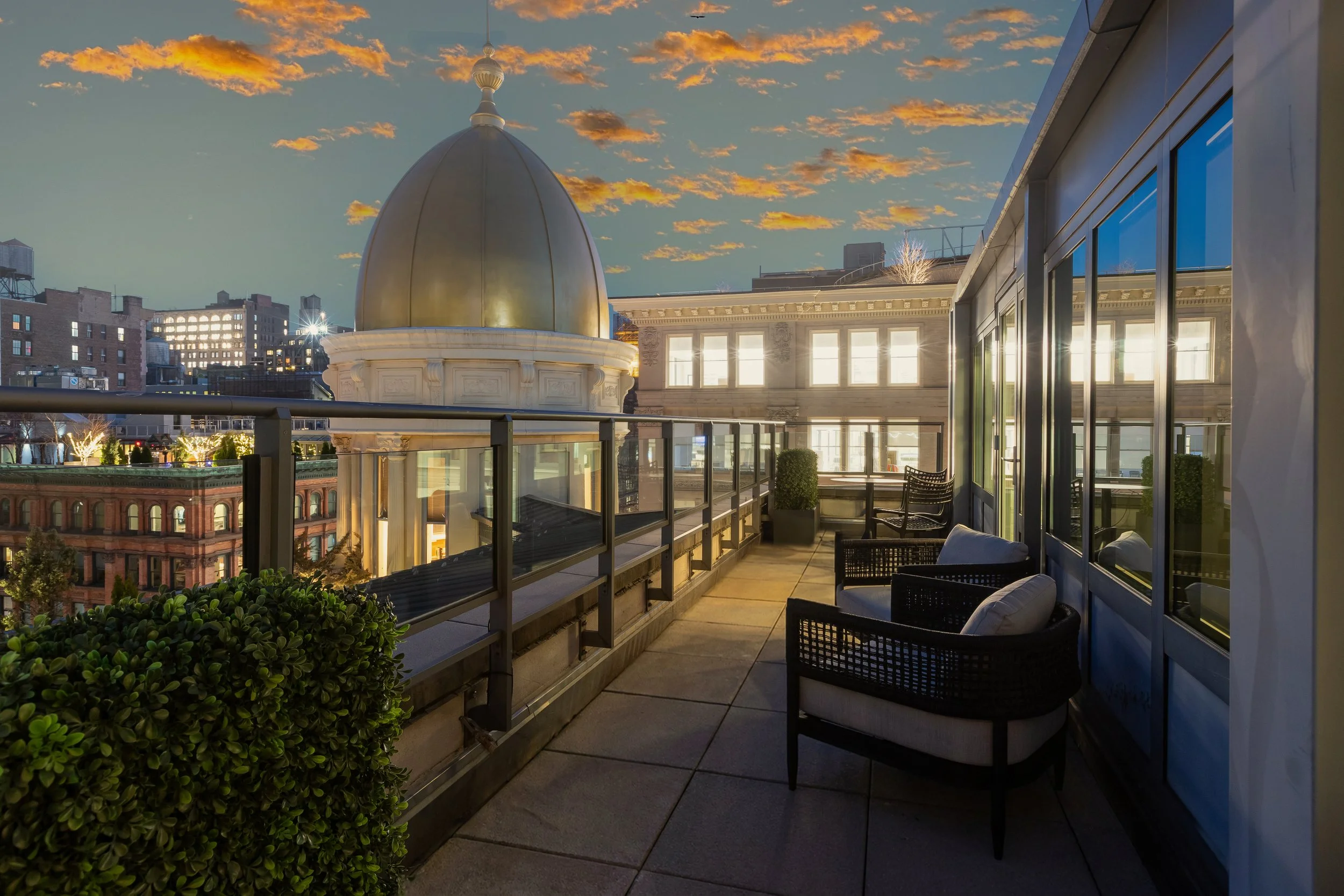 Urban city skyline at sunset from a rooftop balcony with outdoor chairs and potted plants.
